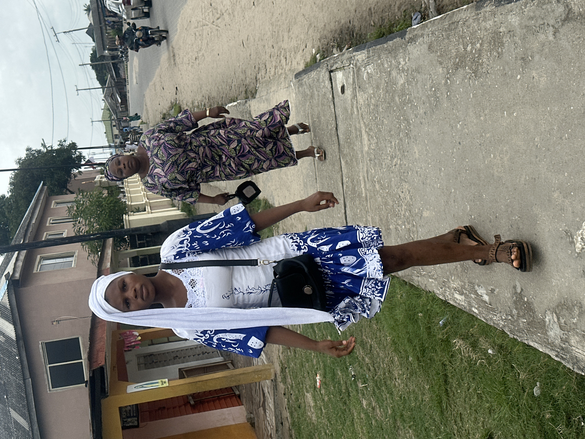 In Badagry, Nigeria, a young Black woman wearing a white headscarf, a white and blue patterned dress, and sandals walks on a concrete sidewalk. She carries a black purse over her shoulder. Behind her, a slightly older Black woman in a purple and green patterned dress also walks on the sidewalk. Further down the street, a motorcycle with a rider is visible, along with other vehicles and buildings. The environment appears to be a street scene in a Nigerian city, with a mix of residential and commercial structures. The sky is overcast.