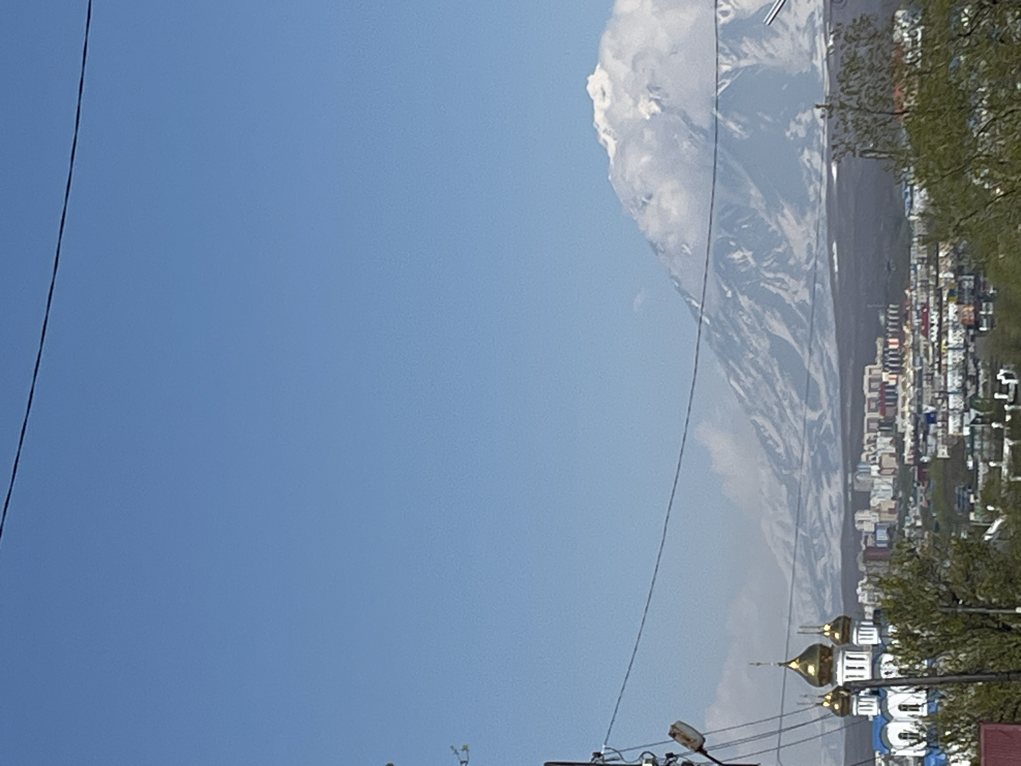 An elevated, daylight panoramic view depicts the city of Petropavlovsk-Kamchatsky, Russia, with a prominent snow-capped volcano in the background. In the foreground on the left, a utility pole with multiple black electrical wires extends diagonally and horizontally across the upper and mid-left portions of the frame. Part of a red roof structure is visible in the bottom left, alongside green tree foliage. The midground features a Russian Orthodox church on the left, characterized by blue walls, white trim, and four golden onion domes, each topped with a cross. To the right and below the church, a densely packed urban residential area spreads across the central midground, composed of numerous multi-story buildings in various muted colors including white, grey, red, and blue. More green tree foliage is interspersed throughout the urban landscape, especially in the lower central and right areas. The background is dominated by a large, snow-covered volcanic mountain range. The most prominent peak, situated in the right background, is heavily snow-covered and partially obscured by white clouds, revealing dark, exposed rock on its lower slopes. The sky above is a clear, uniform light blue. No people, vehicles, or animals are visible, and no actions or interactions are depicted.