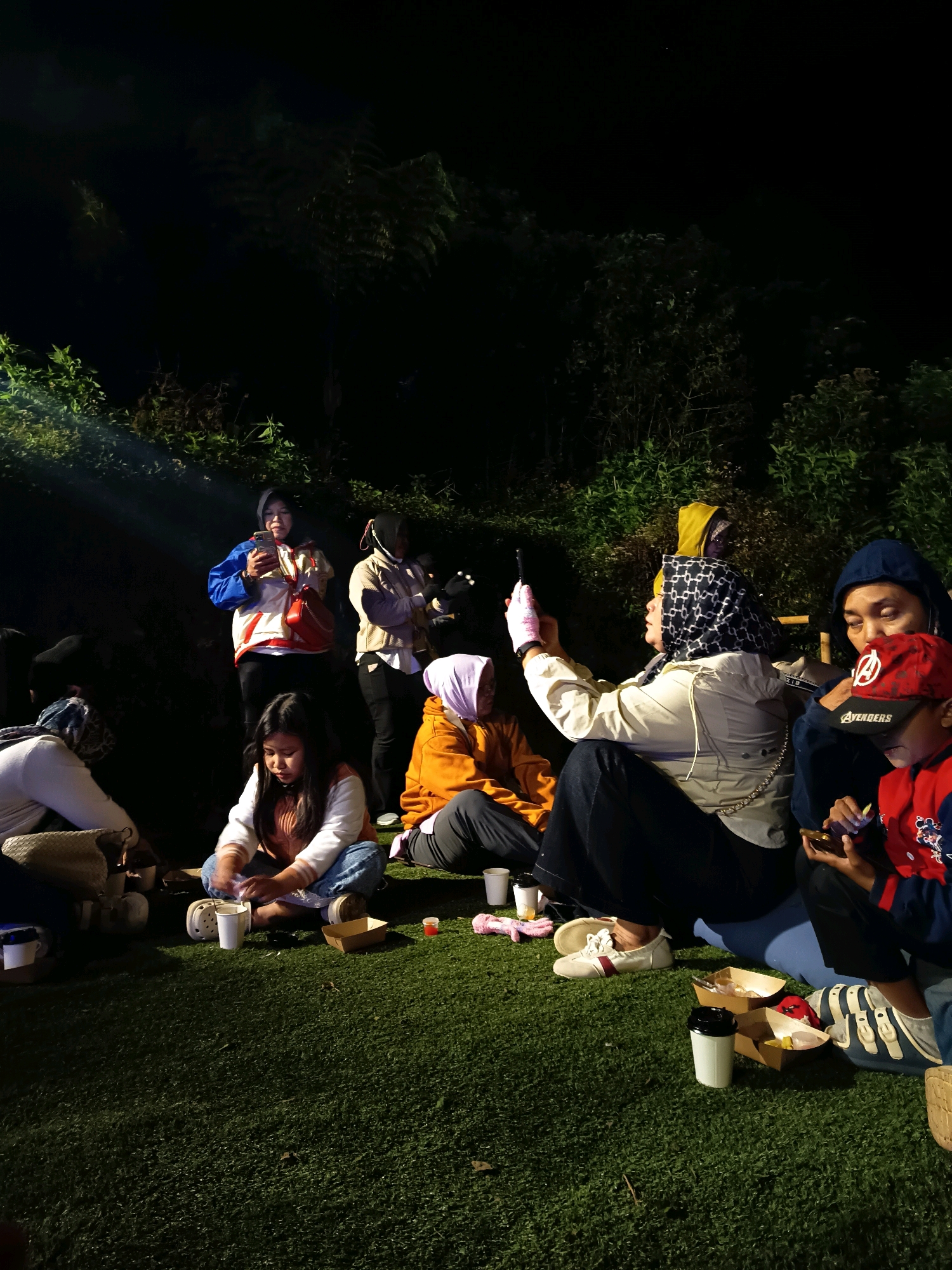 A group of people are gathered outdoors at night, seated on artificial turf in front of dense foliage. The lighting suggests artificial illumination from above, casting shadows and highlighting some individuals. In the foreground, to the right, a young boy wearing a red baseball cap with an "Avengers" logo and a blue and red jacket is looking down at a mobile device. Next to him, an adult in a dark blue hoodie is also looking at a device. To their left, a woman wearing a patterned headscarf and a light-colored jacket is holding up a mobile device, possibly taking a photo or video. A person in an orange hoodie is seated behind her. In the center of the image, a young girl with long dark hair is sitting cross-legged, looking down at something in front of her and holding a cup. To her left, another person in a white top and jeans is seated. Further back, two women are standing. One woman in a blue and red jacket and black pants is holding a mobile device and a red purse. Another woman in a light-colored jacket and black pants is also standing. The environment appears to be a recreational area with a focus on outdoor seating. The presence of food containers and cups suggests a casual gathering. The background is dominated by dark foliage, indicating a natural or semi-natural setting, possibly a park or garden area. The overall scene depicts a social gathering at night, with individuals engaged with their devices and each other.