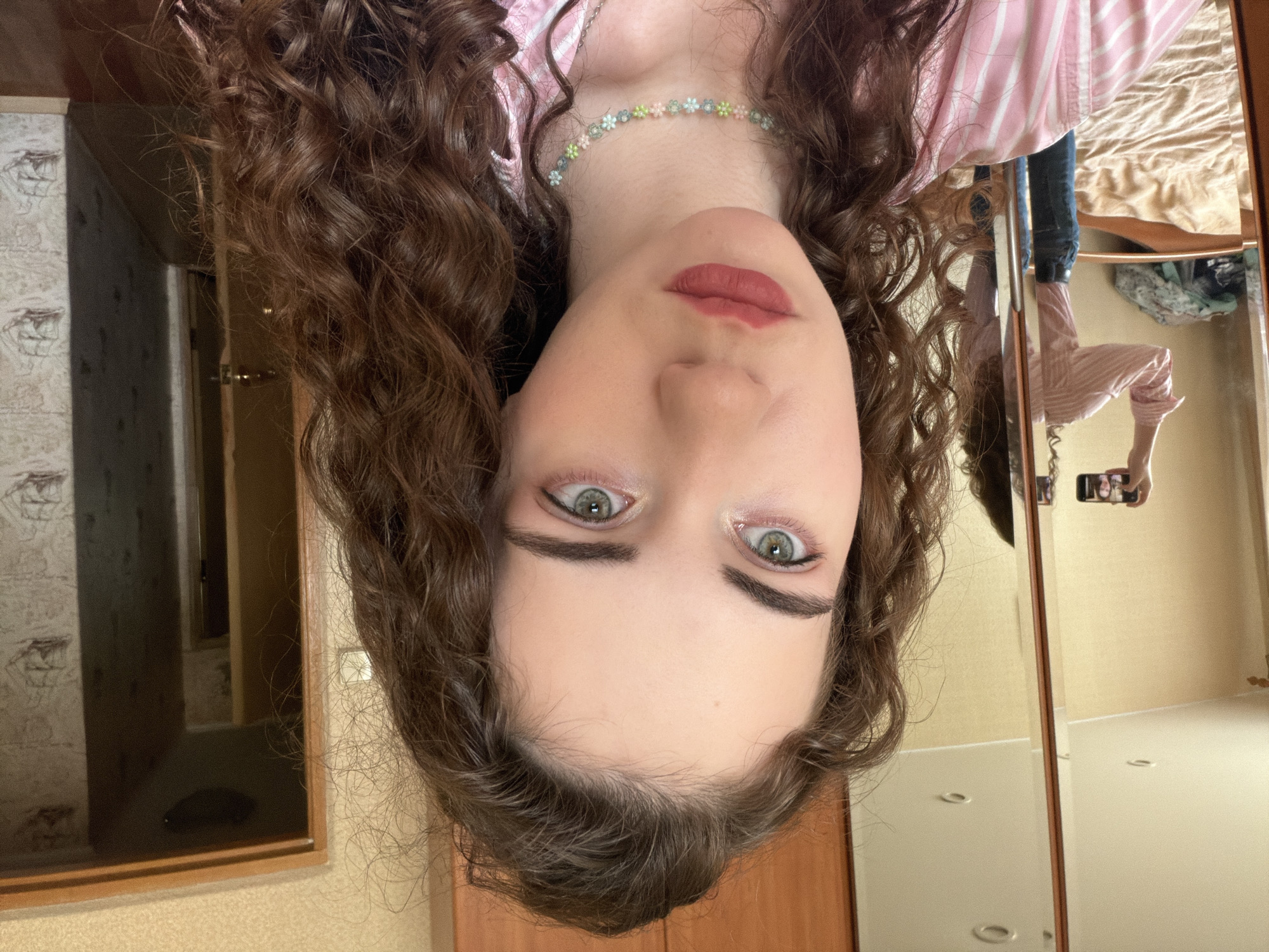 A young woman with long, curly brown hair and light green eyes is positioned in the foreground, looking directly at the camera. She is wearing a pink and white striped shirt and a delicate beaded necklace with floral accents. Her makeup includes matte lipstick in a muted berry shade and subtle eye shadow.
The background reveals a bedroom setting. To the left, a large mirror reflects the woman taking the picture with her phone, as well as a bed with beige bedding and some clothing items on the floor. To the right, a doorway leads into another room, with a glimpse of a hallway. The walls are covered in textured wallpaper with a repeating ship pattern. The overall impression is of a casual, indoor environment.
There is no information in the image that suggests the location is in the city of Zavodoukovsk, Russia.