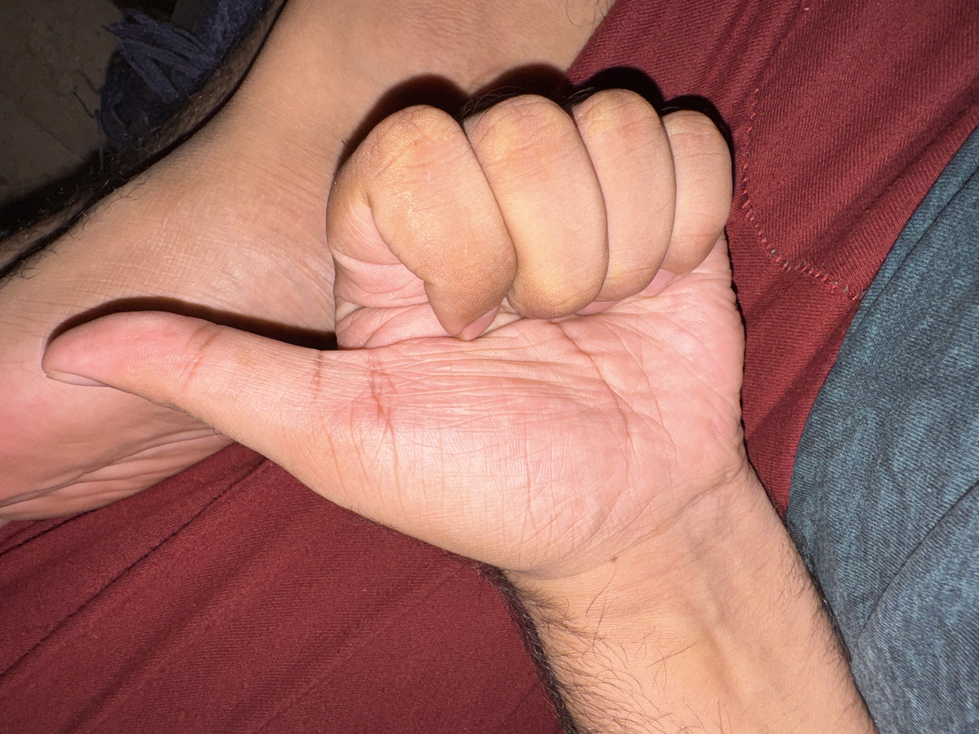 A close-up shot shows a person's right hand clenched in a fist with the thumb extended, positioned on a surface comprised of a maroon fabric and a blue-gray fabric. The skin on the hand is light brown with prominent lines and wrinkles. Hair is visible on the wrist and lower forearm. A partially visible foot is present in the upper left corner. The location is Unknown, Unknown.