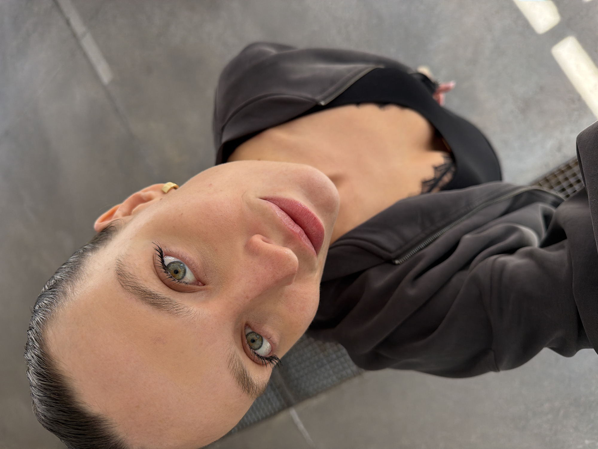 A close-up, rotated portrait features a woman looking directly at the camera from a low angle. Her head is positioned towards the bottom-left of the frame, with her body extending diagonally upwards. She has fair skin and striking green/hazel eyes with dark, full eyelashes. Her eyebrows are well-defined, and her lips, colored in a soft pink, are slightly parted. Her hair is styled back and appears sleek, revealing her hairline. She is wearing a dark hooded jacket, partially unzipped to show a black lace-trimmed top or bra underneath. A small golden ear cuff or earring adorns her left ear. The background is a grey, textured surface, possibly concrete, with some lighter reflections suggesting ambient light. There is no visible text. The woman's expression is neutral yet engaging, captured in what appears to be a selfie or a close-up portrait. The provided context states the location is Budva, Montenegro, although no specific landmarks are visible in the image.