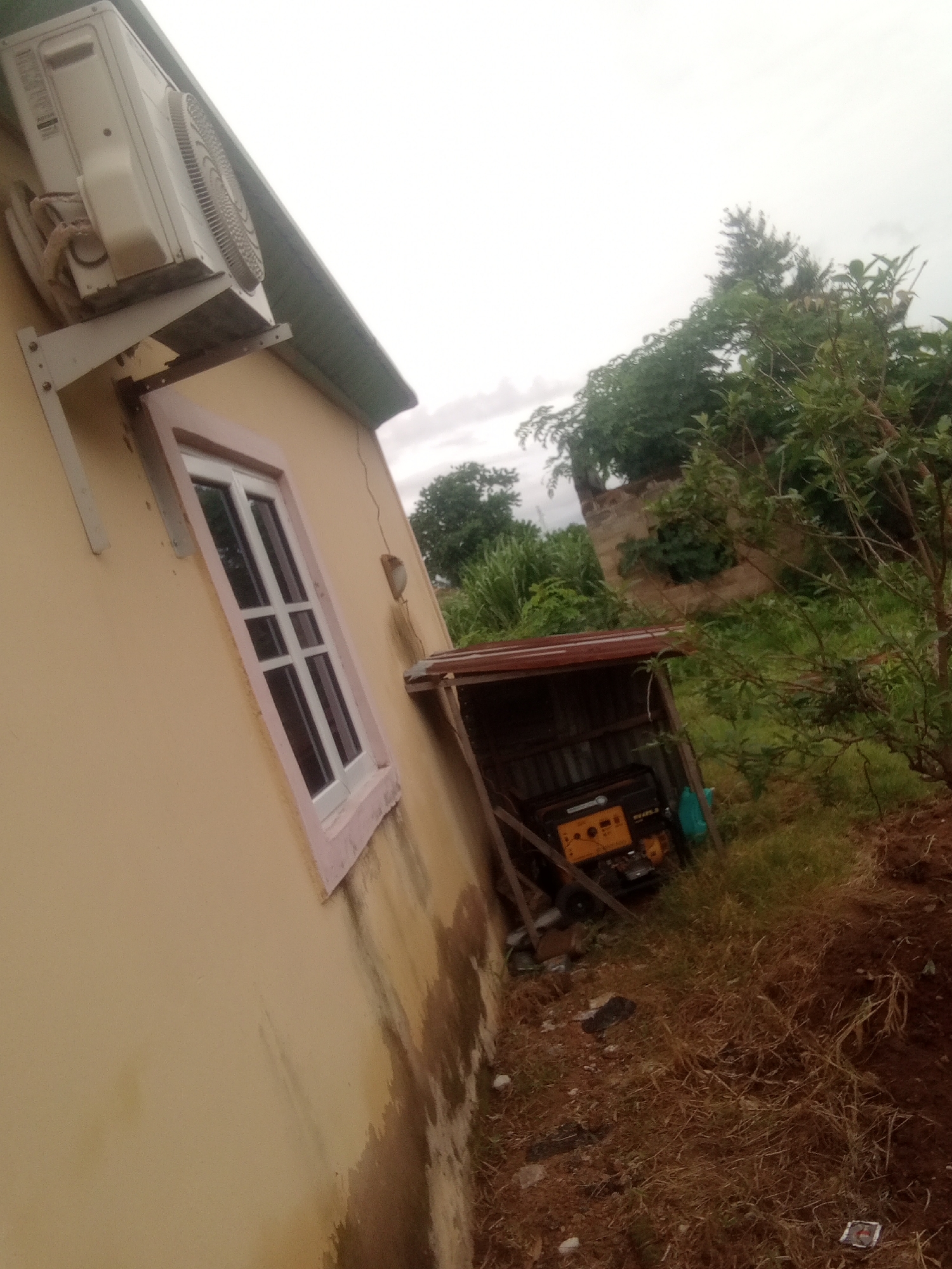 The image shows an outdoor scene of a residential building with an air conditioning unit mounted on the wall. A window with a pink frame is visible on the side of the building. Adjacent to the building, there is a makeshift shelter covered with corrugated metal, under which a yellow generator is placed. The surrounding area is overgrown with grass and some trees, indicating a somewhat rural or unkempt setting. The sky is overcast, suggesting a cloudy day. There are no people visible in the image. The location is Jalingo, Nigeria. The generator has text that appears to read "EVEREST 4500".