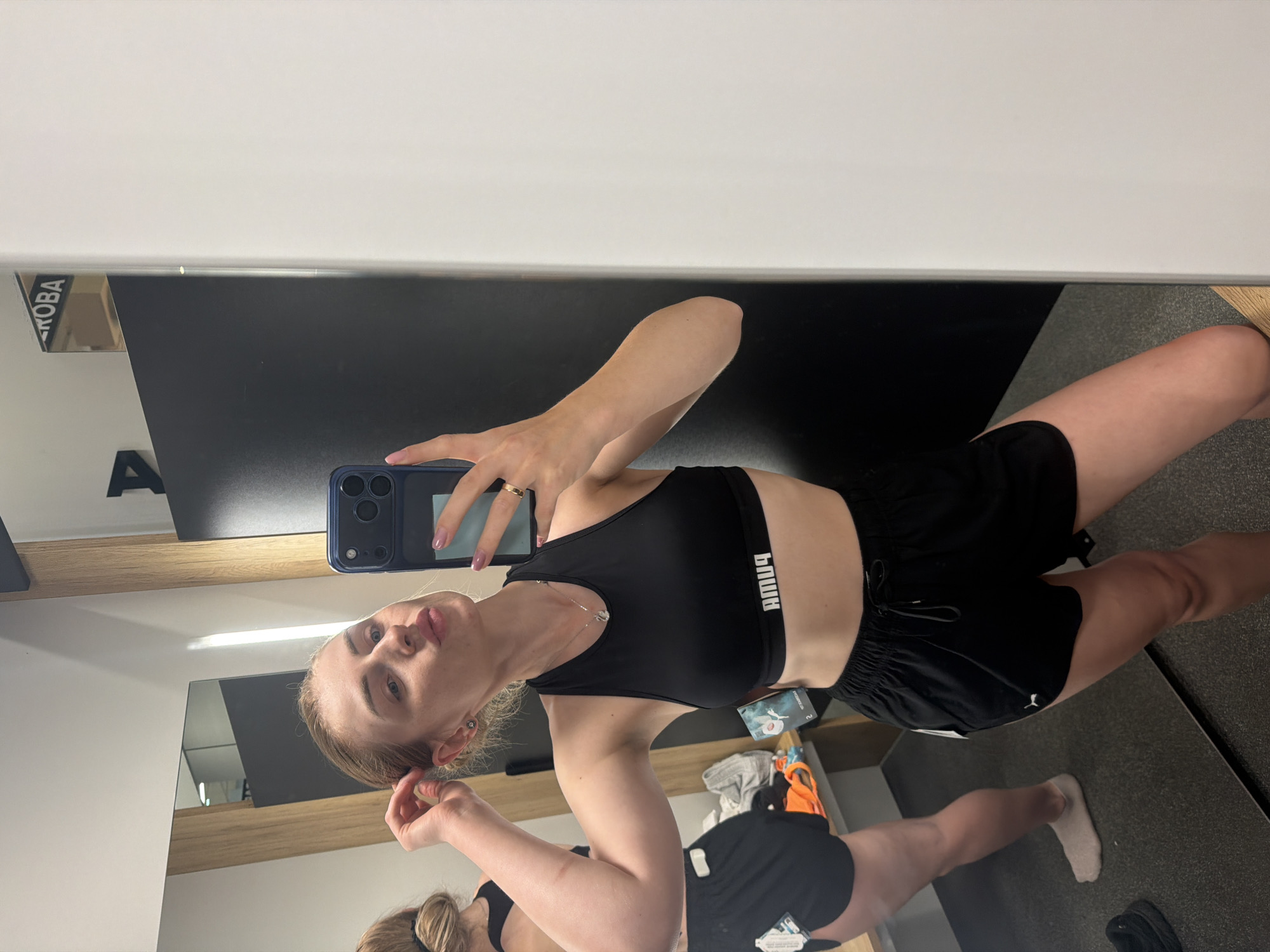 A young woman with blonde hair tied back is taking a mirror selfie in what appears to be a fitting room or changing area. She is wearing a black Puma sports bra and black Puma athletic shorts. Her left hand is touching her hair near her temple, and her right hand holds a dark blue smartphone in front of her face. She is puckering her lips. The background shows a mirror reflecting another person in similar athletic wear, a wooden changing room structure, and a black partition. The floor is dark gray. There are some folded clothes and tags visible on a shelf in the reflection.