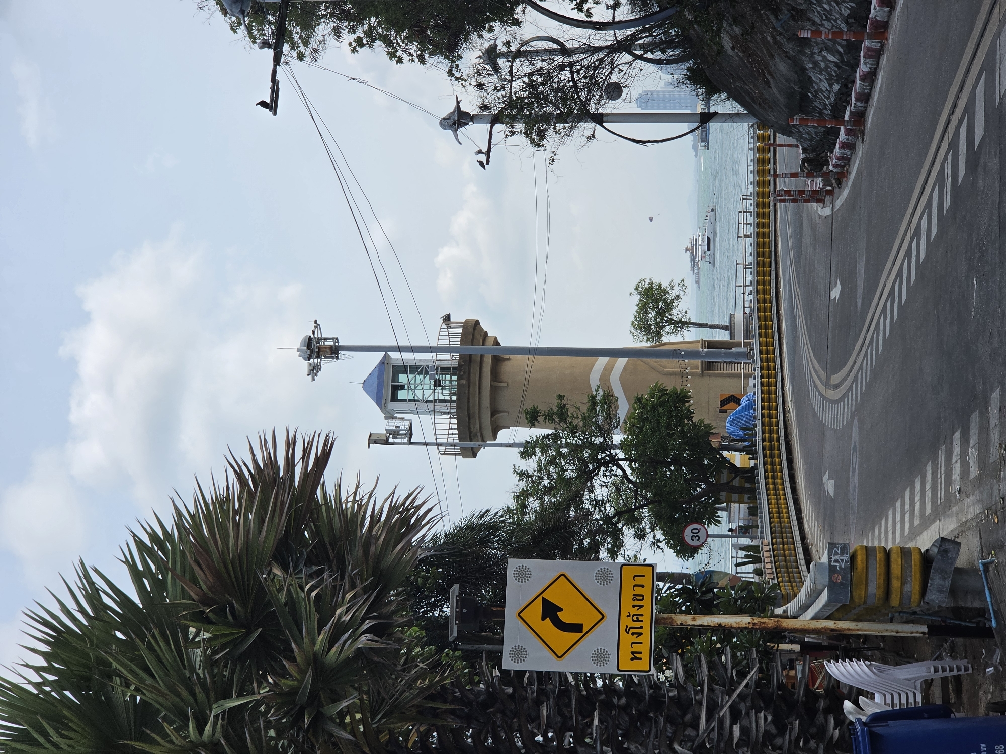 The image captures a coastal scene, likely in Pattaya City, Thailand, during the daytime with clear weather and scattered white clouds.
In the foreground, on the bottom left, are dense green palm-like plants with spiky leaves. A prominent yellow diamond-shaped sign with a black border displays an arrow curving to the left, indicating a left curve in the road. Below this, a white rectangular sign has Thai text "ทางโค้งซ้าย" (Thang Khong Sai), meaning "Left Curve".
The mid-ground features a paved road with yellow double lines and white dashed markings, curving to the right from the bottom right towards the top right of the frame. A white arrow on the road directs traffic. A yellow and black barrier, along with some orange traffic cones, separates the road from the sea on the left. Behind this barrier, a concrete wall with a visible rocky texture rises. A red circular sign with "30" on it, likely a speed limit, is visible near the road. Dominating the left side of the mid-ground is a light brown tower, possibly a lighthouse or observation post, featuring a blue-domed cap and various communication antennas. It is partially obscured by lush green trees.
The background reveals the expanse of the sea, with several boats dotting the water, including a larger white vessel. A small white shape in the distance above the sea suggests a paraglider. Distant buildings or a cityscape are faintly visible on the horizon.
Several utility poles with a dense network of overhead wires crisscross the upper portion of the sky. A person wearing a blue top is partially visible near the road on the right, behind some vegetation. White plastic chairs and a blue waste bin are seen in the bottom right corner. The overall scene depicts a functional coastal road with a scenic view, implying both local infrastructure and recreational opportunities.