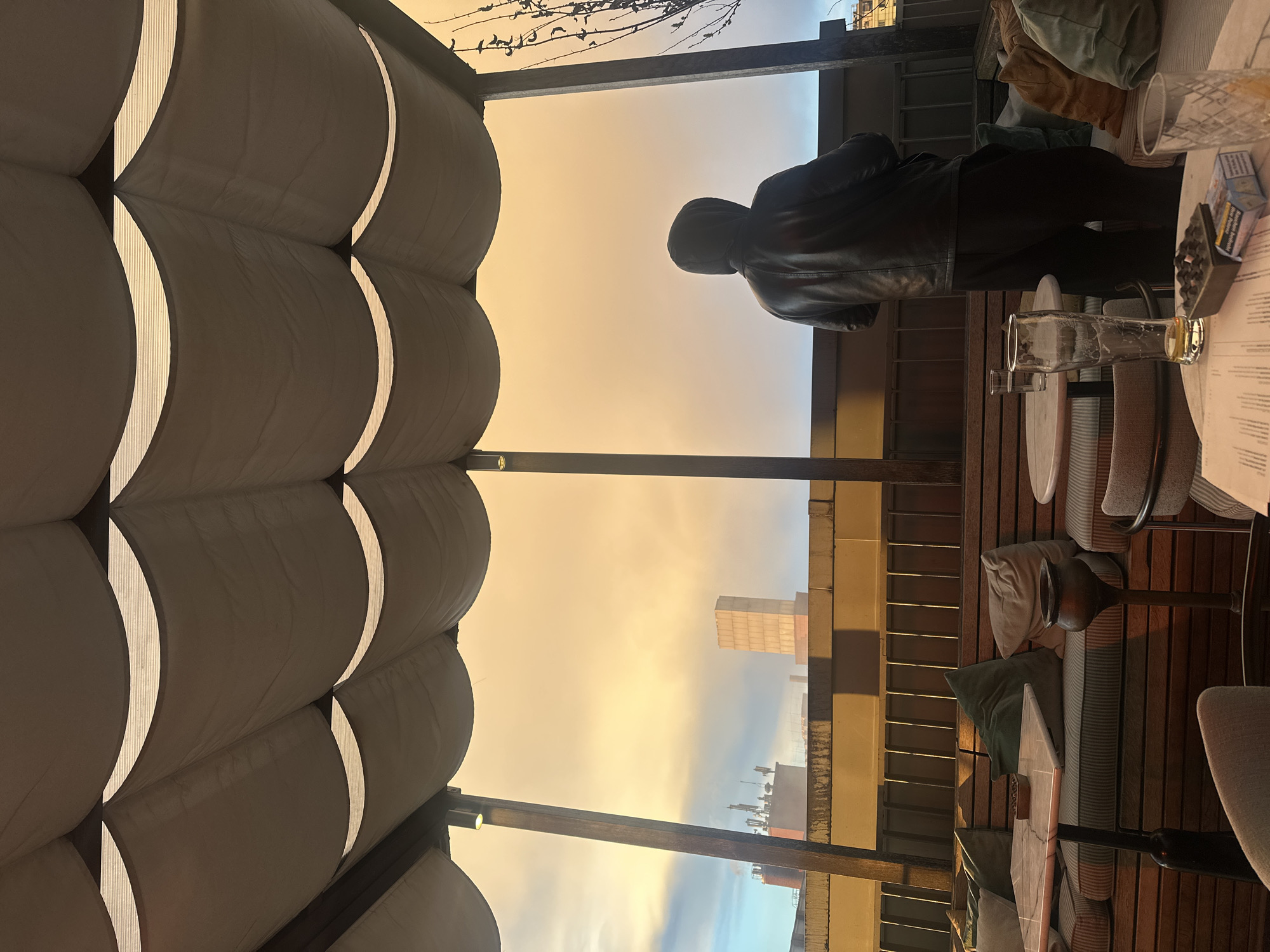 The image depicts a person in a black jacket standing on a balcony overlooking a cityscape under an orange and grey sky, likely at sunset or sunrise. The scene is framed by a ribbed, white awning above, and a wooden structure that supports the balcony. On the right side of the frame, there is a table with glassware, an ashtray, cigarettes, and papers. Beyond the balcony, buildings can be seen in the distance. Given the architectural style of the buildings in the background, the photo was likely taken in Berlin, Germany.