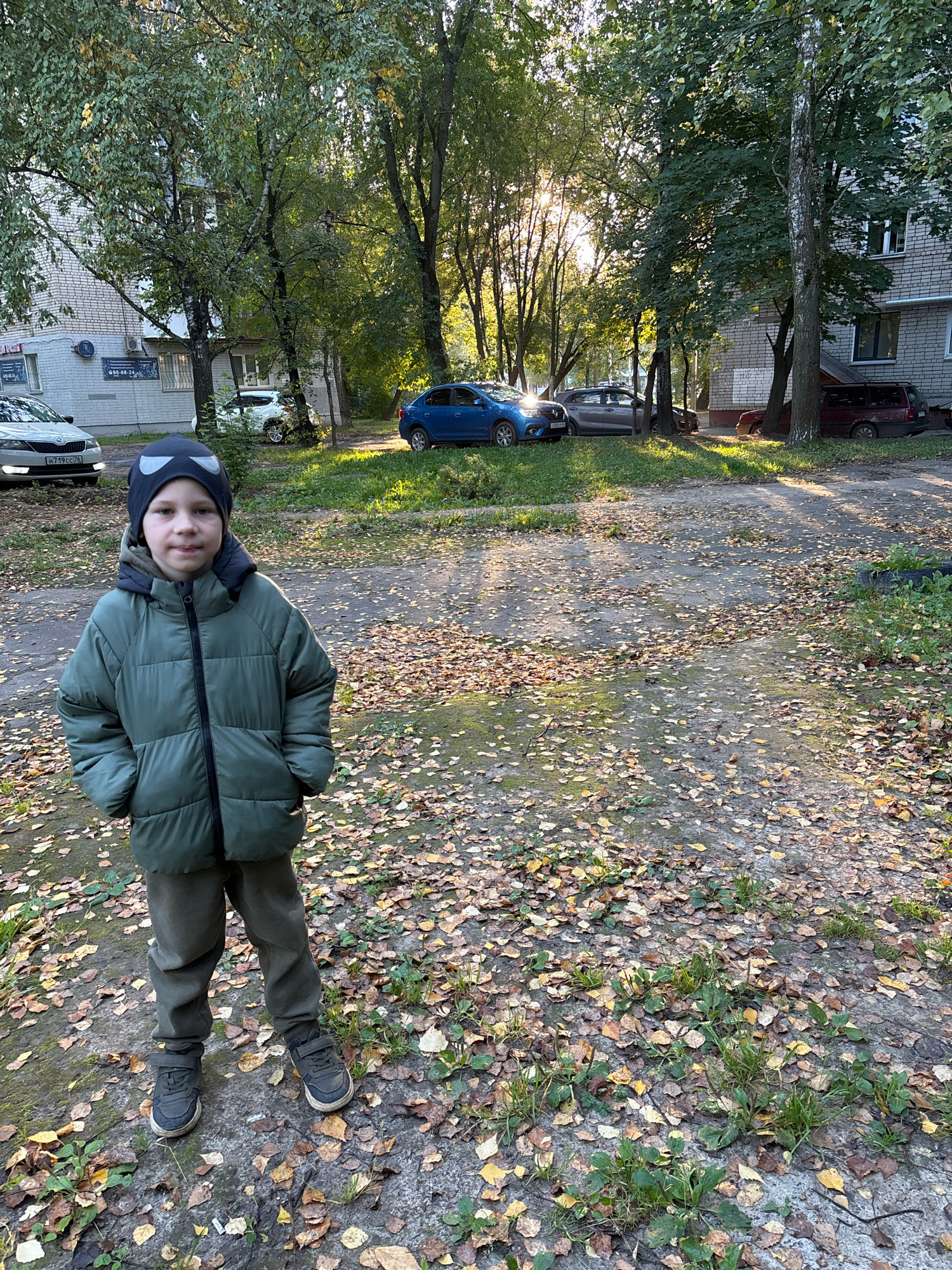 A young boy stands in the foreground of a courtyard in Rusanovo, Russia. He is wearing a green puffer jacket, grey pants, and dark sneakers, and a black beanie with white eye-like patterns. The boy has his hands in his pockets and is looking at the camera. The ground around him is covered in fallen leaves, indicating it is autumn. In the background, several cars are parked on a paved area. Behind the cars, there is a brick apartment building and tall trees with green and yellow leaves. Sunlight is filtering through the trees, creating dappled light on the ground. The overall scene depicts a typical residential courtyard in Russia during the fall season.