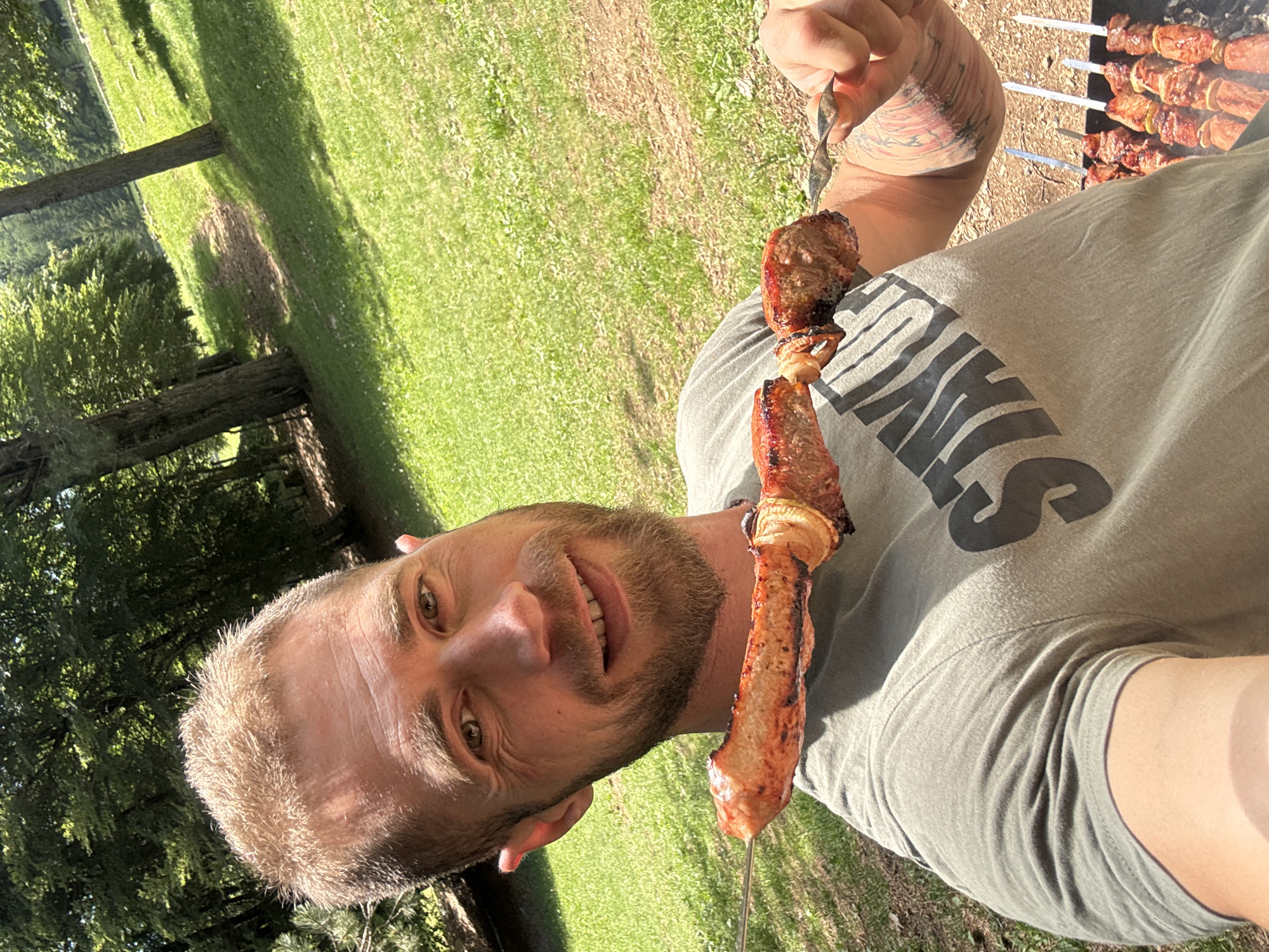 A male individual is depicted in a close-up, outdoor selfie, situated in Dolovi, Montenegro. The subject has short, spiky blonde hair and a light brown beard and mustache, smiling directly into the camera. He wears an olive green t-shirt with the upper portion of dark lettering, appearing to read "STAY...", visible on his chest. His left forearm displays a multi-colored tattoo. He holds a metal skewer horizontally with his right hand, presenting several pieces of grilled meat, consistent with shashlik or kebab, interspersed with sliced onions. The meat pieces exhibit a reddish-brown, slightly charred exterior. In the lower right quadrant of the image, a charcoal grill (mangal) is visible on the ground, supporting multiple additional metal skewers loaded with similar grilled meat and onions, with subtle indications of rising heat or smoke. The immediate surroundings consist of green grass and dried earth. The background features a sloping, grassy area with a visible dirt path ascending uphill, flanked by mature deciduous trees under bright sunlight, which casts distinct shadows. The scene is illuminated by strong natural daylight, suggesting a clear, sunny outdoor environment.