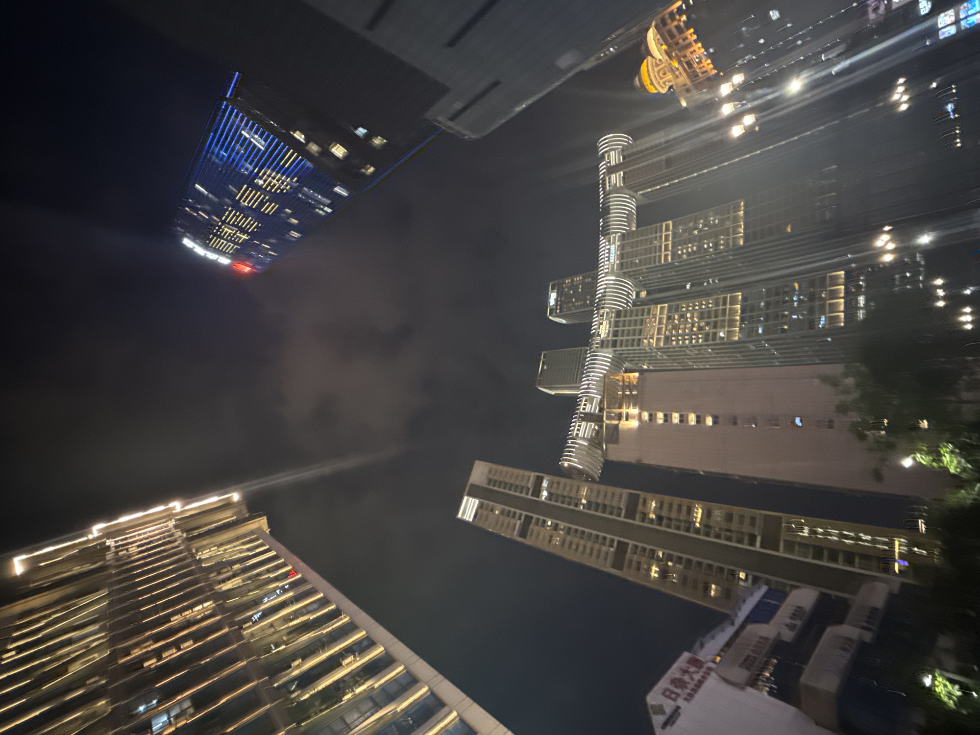The image captures a nighttime cityscape in the Yuzhong District, China, characterized by a dramatic low-angle perspective looking upwards at illuminated skyscrapers. The architecture is modern and diverse, with various building styles visible. On the left, a tall, slender building with a façade of vertical golden lights dominates the upper portion of the frame. To its right, a prominent skyscraper features blue and yellow lighting, with some windows illuminated. In the mid-ground, several interconnected skyscrapers are visible, featuring a distinctive design with curved, illuminated elements connecting them. These structures also have many lit windows. Further to the right, a building with a golden dome and illuminated windows is partially visible. The sky is dark and cloudy, with light pollution from the city creating a hazy glow. The foreground shows the tops of trees and streetlights, suggesting a street-level view looking up. The overall impression is one of a vibrant, modern urban environment at night.