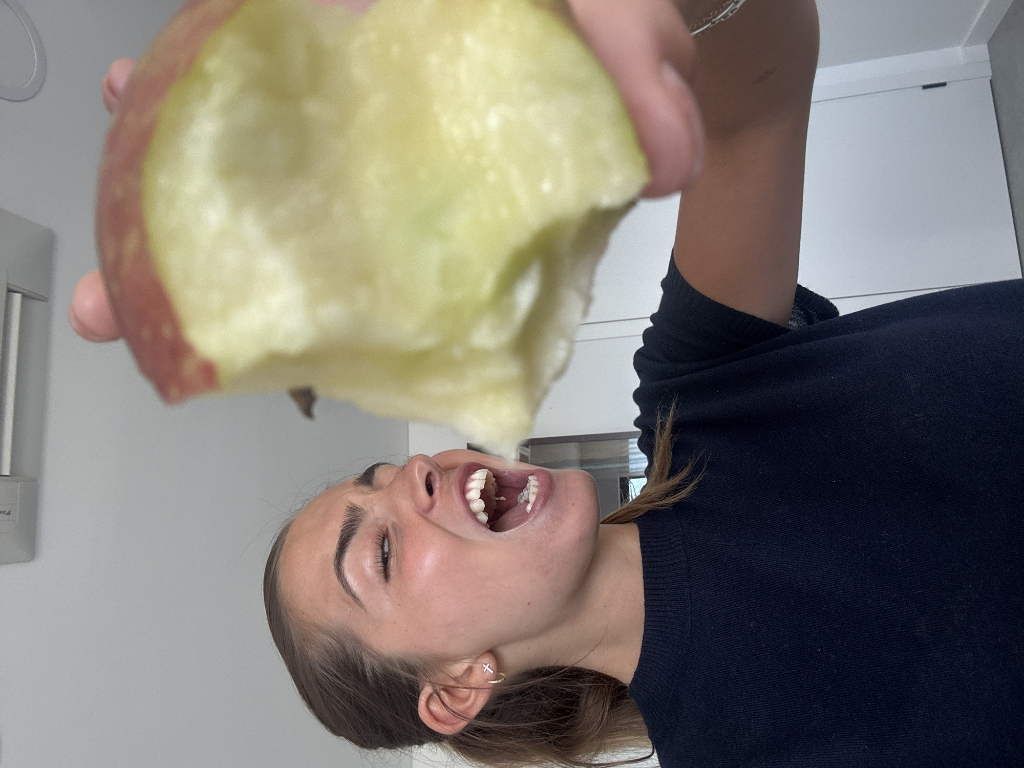 A woman is holding a partially eaten apple above her open mouth. She is wearing a dark blue sweater and earrings. The background is a white wall and a blurry window. The photograph appears to be taken indoors. The location in the city of Torres Vedras, Portugal is not discernible from the image.