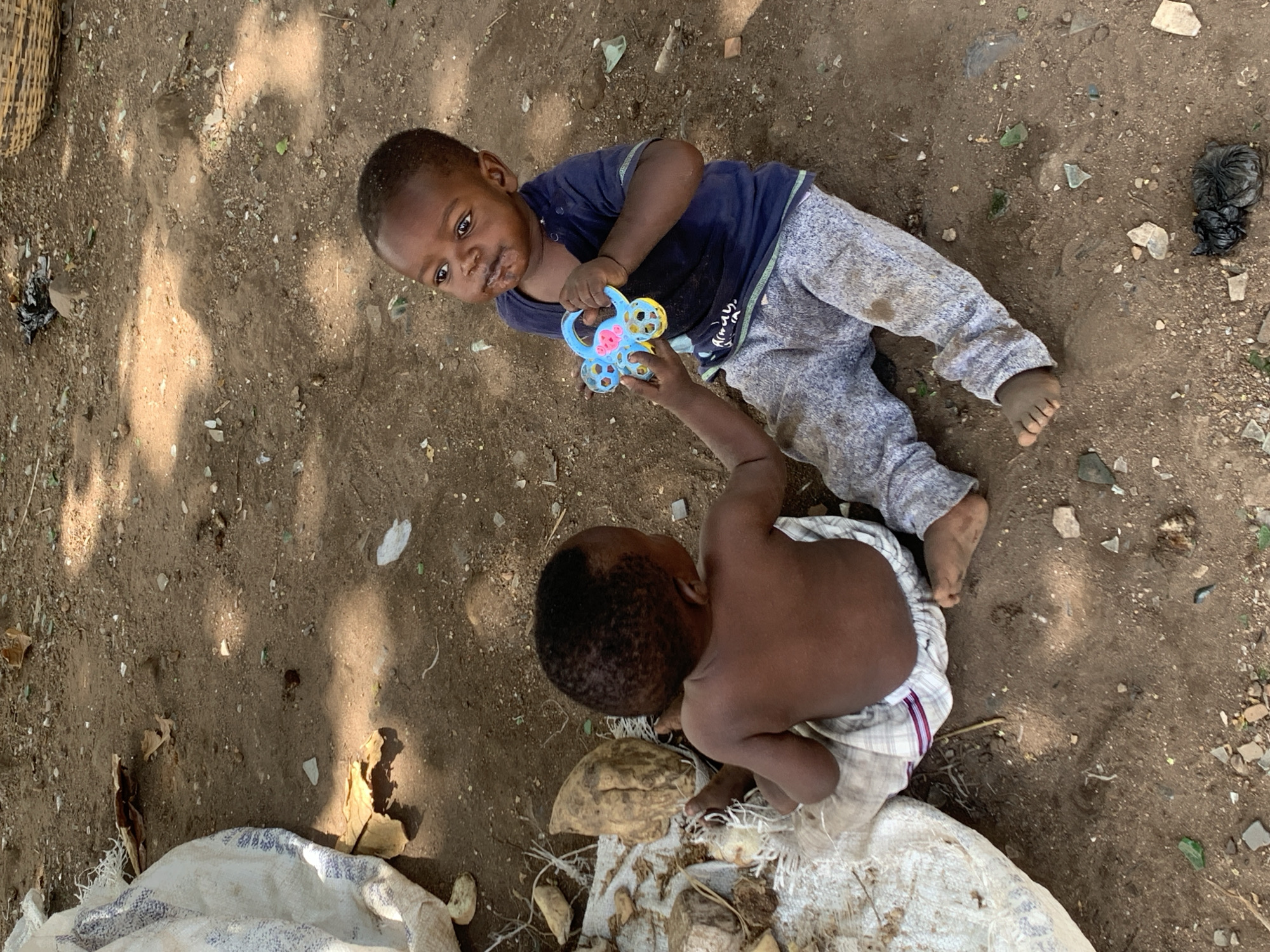 An overhead photograph captures two young children on a sun-dappled dirt ground, likely in an outdoor setting in Ilorin, Nigeria. The scene is illuminated by strong sunlight filtering through an unseen canopy, casting dappled shadows across the earth.
In the foreground, a child with a lighter complexion is lying on their side, wearing a dark blue t-shirt and light grey pants. This child looks towards the viewer with a neutral expression, holding a brightly colored plastic toy – blue with yellow and pink elements – in their hands. Faint, possibly incomplete text resembling "ALVAH" can be seen on the grey pants.
Beside them, a second child with a darker complexion is positioned on their knees or squatting. This child is shirtless, clad in a light-colored, possibly striped fabric wrapped around their lower body. Their face is obscured as they lean forward, reaching out towards the toy held by the first child, suggesting a moment of shared play or interaction.
The ground is a mixture of dry dirt and small pebbles, littered with natural debris like dry leaves and scattered fragments of glass or other discarded materials. A crumpled black plastic bag is visible in the upper right. In the upper left, a portion of a woven basket is present, while a white sack with blue patterns, holding what appears to be root vegetables, is visible in the lower left. The overall impression is of children playing amidst simple, everyday surroundings.