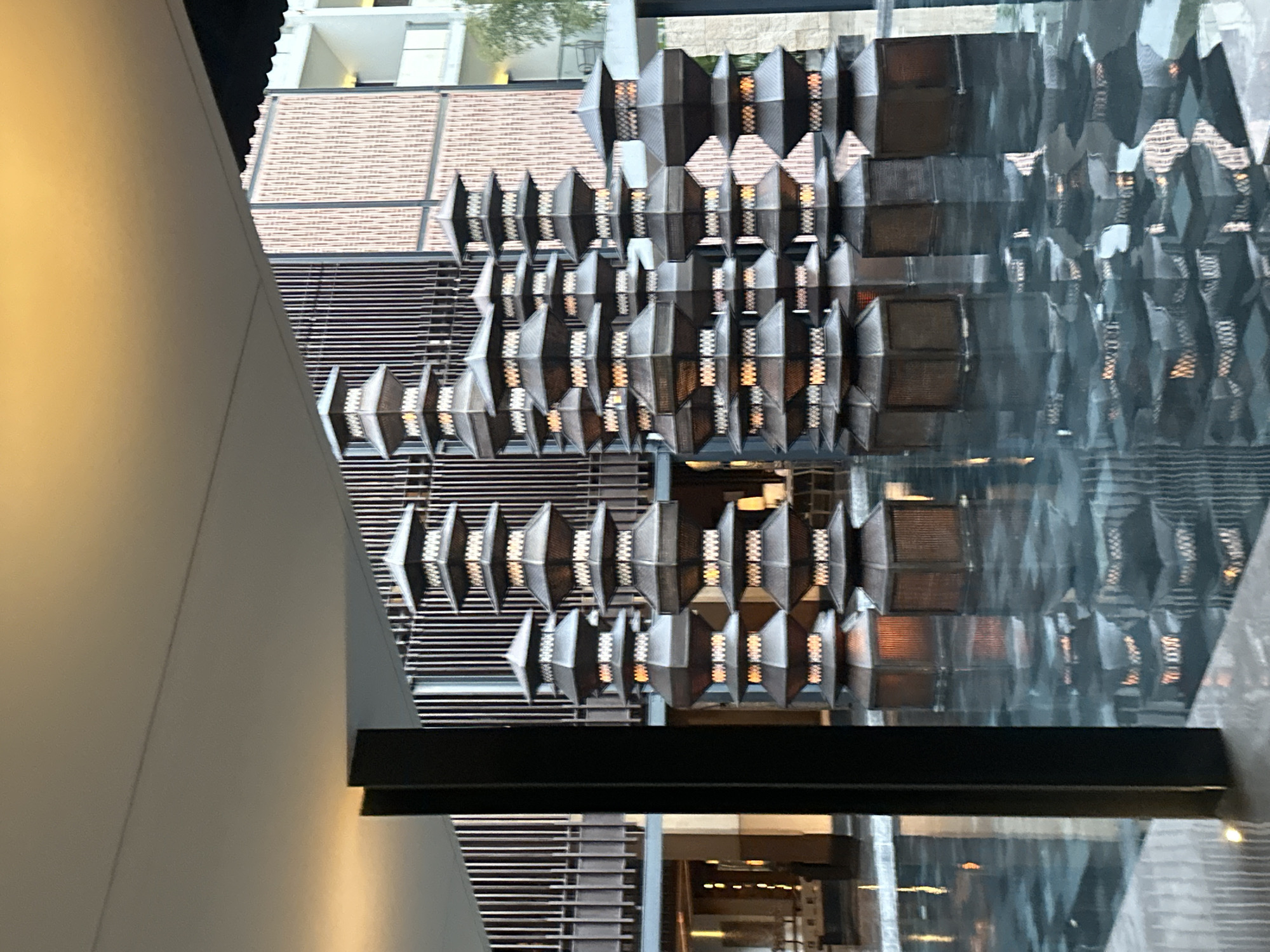 The image displays an interior setting with a striking display of hanging lanterns. The lanterns are geometric in shape, with metallic frames and woven or textured inserts that allow light to emanate from within, creating a warm glow. They are arranged in rows and columns, forming a visually complex and decorative element.
In the background, architectural features are visible, including what appears to be a building with textured brickwork and windows. There are also horizontal slatted elements, possibly part of a wall or decorative screen, and a reflective surface that mirrors the lanterns and surrounding environment, adding depth to the scene. The lighting suggests it is either daytime or a well-lit indoor space.
The scene is set in Kuta, Indonesia. The overall impression is one of sophisticated interior design, with a focus on decorative lighting and architectural interest. No people are visible in the frame, and no text is discernible. The image appears to be a still photograph captured from a slightly elevated perspective, looking down and across the display.