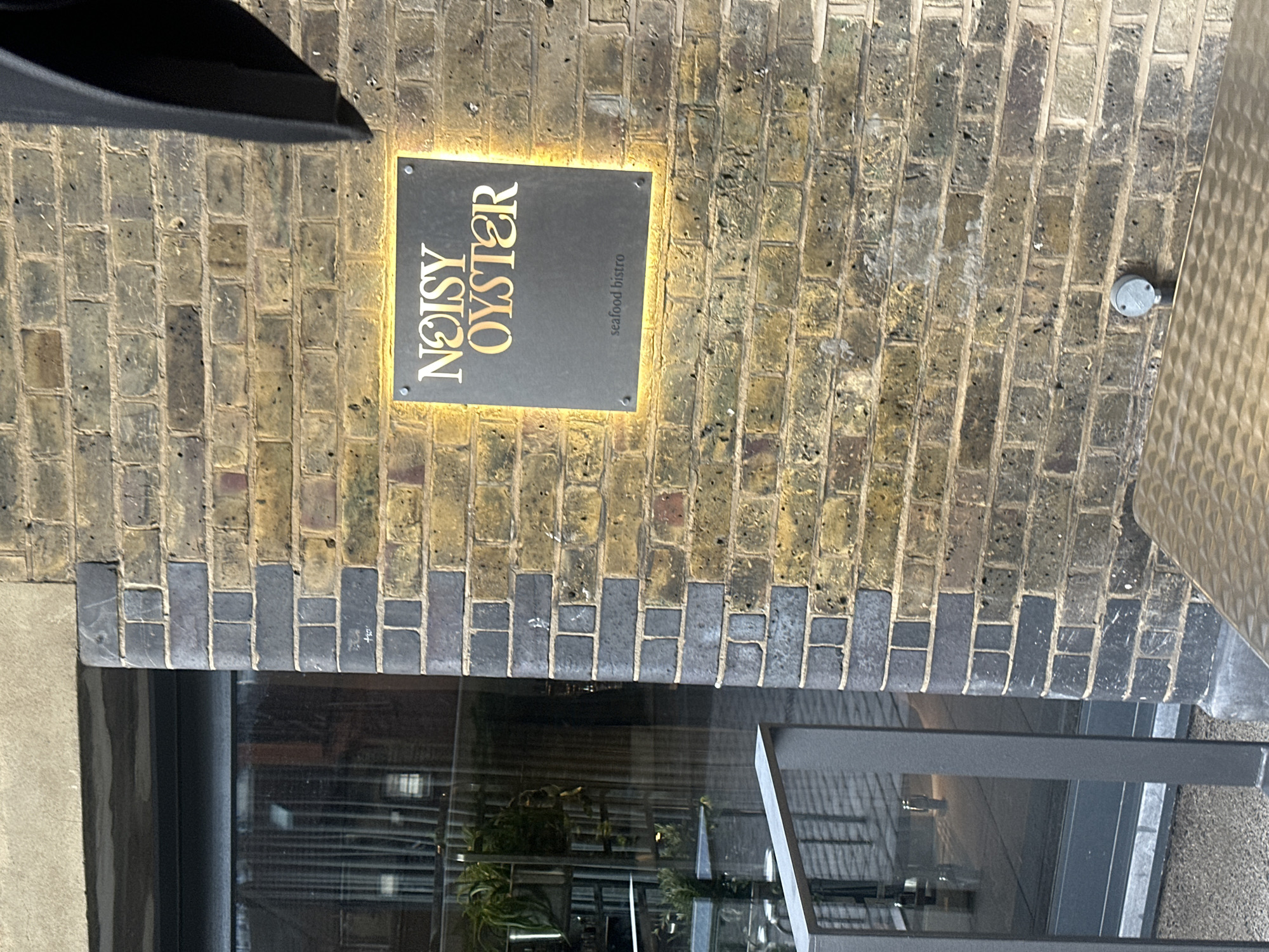 The image shows the exterior of a restaurant named "Noisy Oyster" in London, United Kingdom. The restaurant's sign is mounted on a brick wall. The sign is black with gold lettering, illuminated by a backlight. The words "Noisy Oyster" are written in a vertical stack, and below that, in smaller font, are the words "seafood bistro." Below, there is a modern architectural structure. The brickwork above the window is composed of yellow and grey bricks. A grey electrical box is mounted on the wall to the right of the sign. The image is taken during the daytime. The building appears to be made of brick.