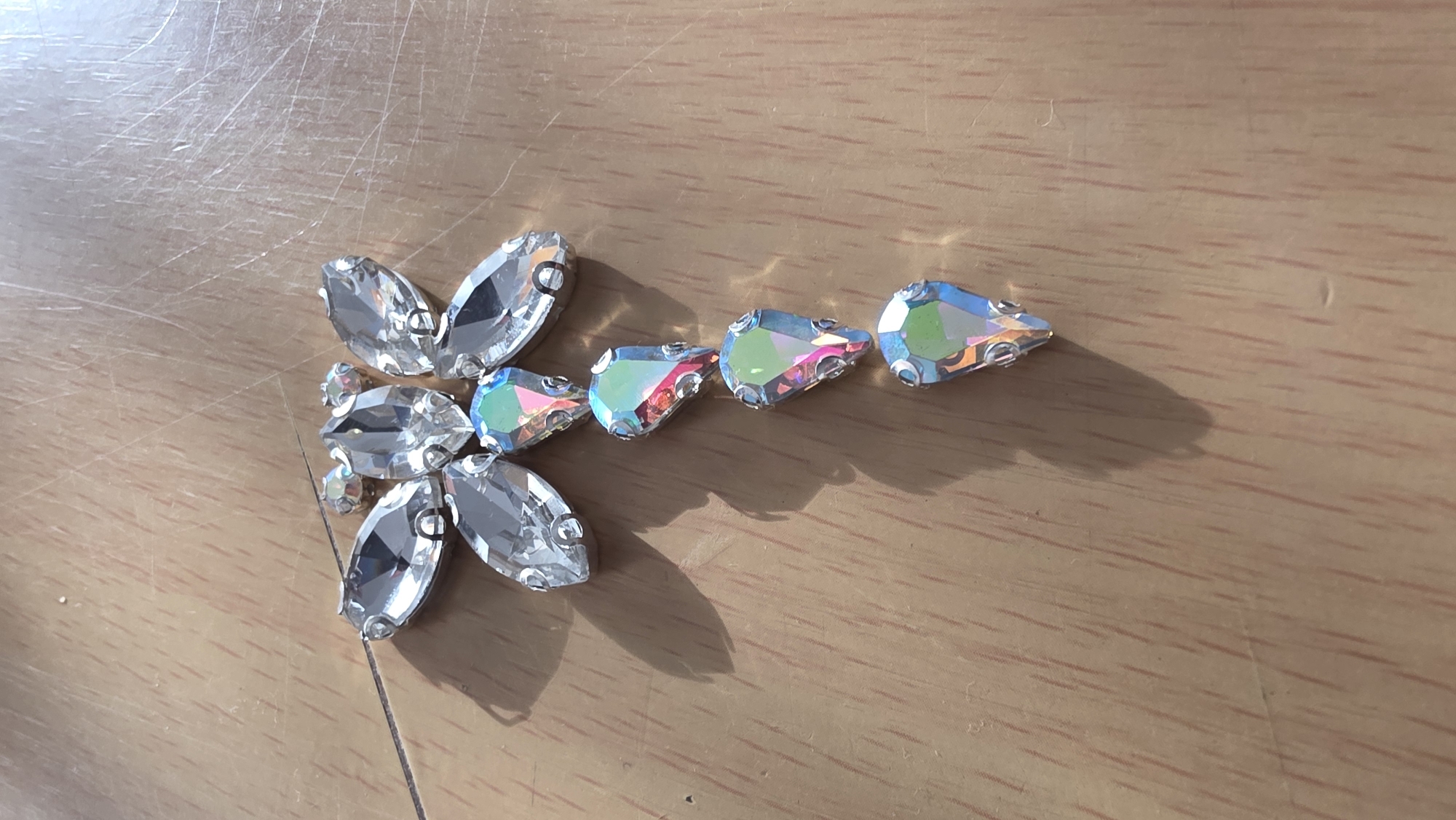 The image shows a close-up of several decorative gems or rhinestones arranged on a wooden surface. Some are clustered in a flower-like shape, while others are lined up in a row. The gems are reflective and appear to be clear or iridescent, with a metallic backing visible around the edges. The wooden surface is light brown with visible grain and some scratches. The location in the city of Blagoevgrad, Bulgaria, cannot be determined from the visual information provided.