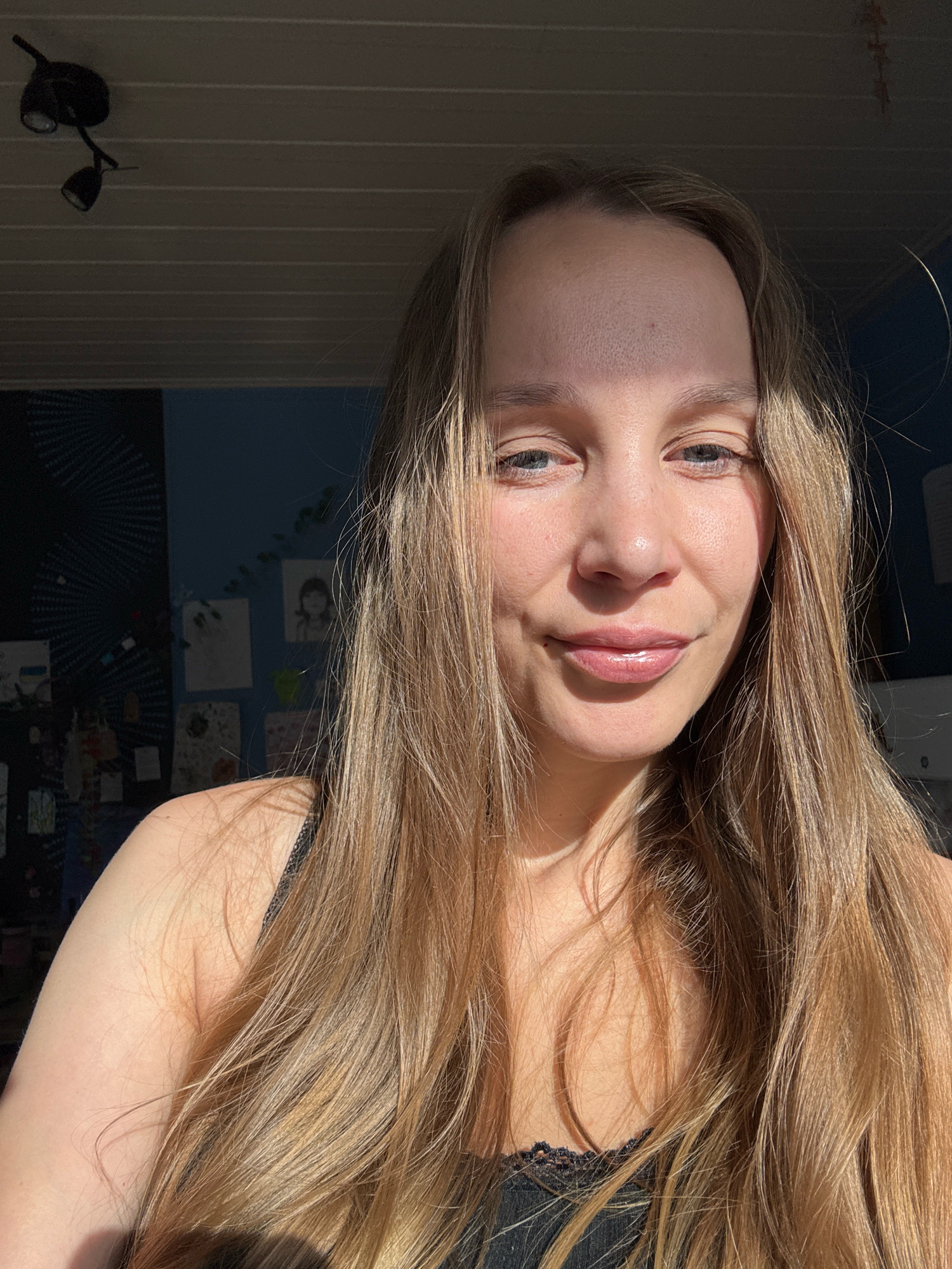The image shows a medium shot of a young woman with long, light brown hair. She is looking at the camera with a slight smile and her eyes are blue. The lighting suggests she is outdoors or near a window with sunlight hitting her face. In the background, there are some faint posters or pictures on a blue wall, and a ceiling light fixture. The overall impression is a casual, natural portrait. The image does not provide any specific information about the location in the city of Überherrn, Germany.