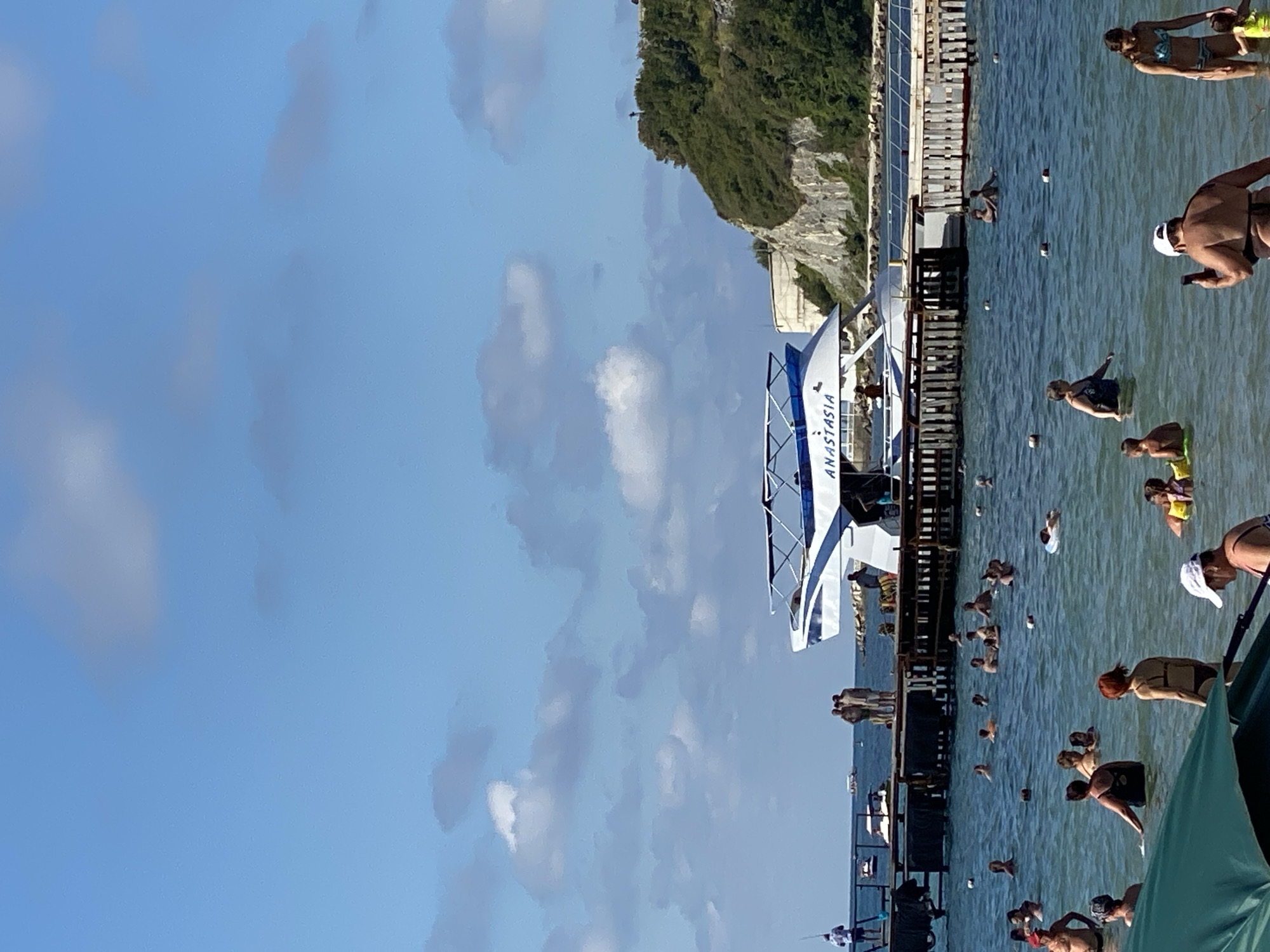A wide shot captures a bustling beach scene in Arkhipo-Osipovka, Russia, on a clear day with a bright blue sky dotted with white clouds. In the foreground, several people are wading in the calm, shallow water. Some are swimming, while others stand and converse. Further out, a long wooden pier juts into the sea, lined with small buoys. At the end of the pier, a modern white and blue speedboat named "ANASTASIA" is docked. A group of people stand on the pier, some appearing to be preparing to board the boat. In the background, a lush green hillside with rocky outcrops rises above the pier and the sea, characteristic of the coastal landscape of Arkhipo-Osipovka. The overall atmosphere is one of leisure and summer activity.