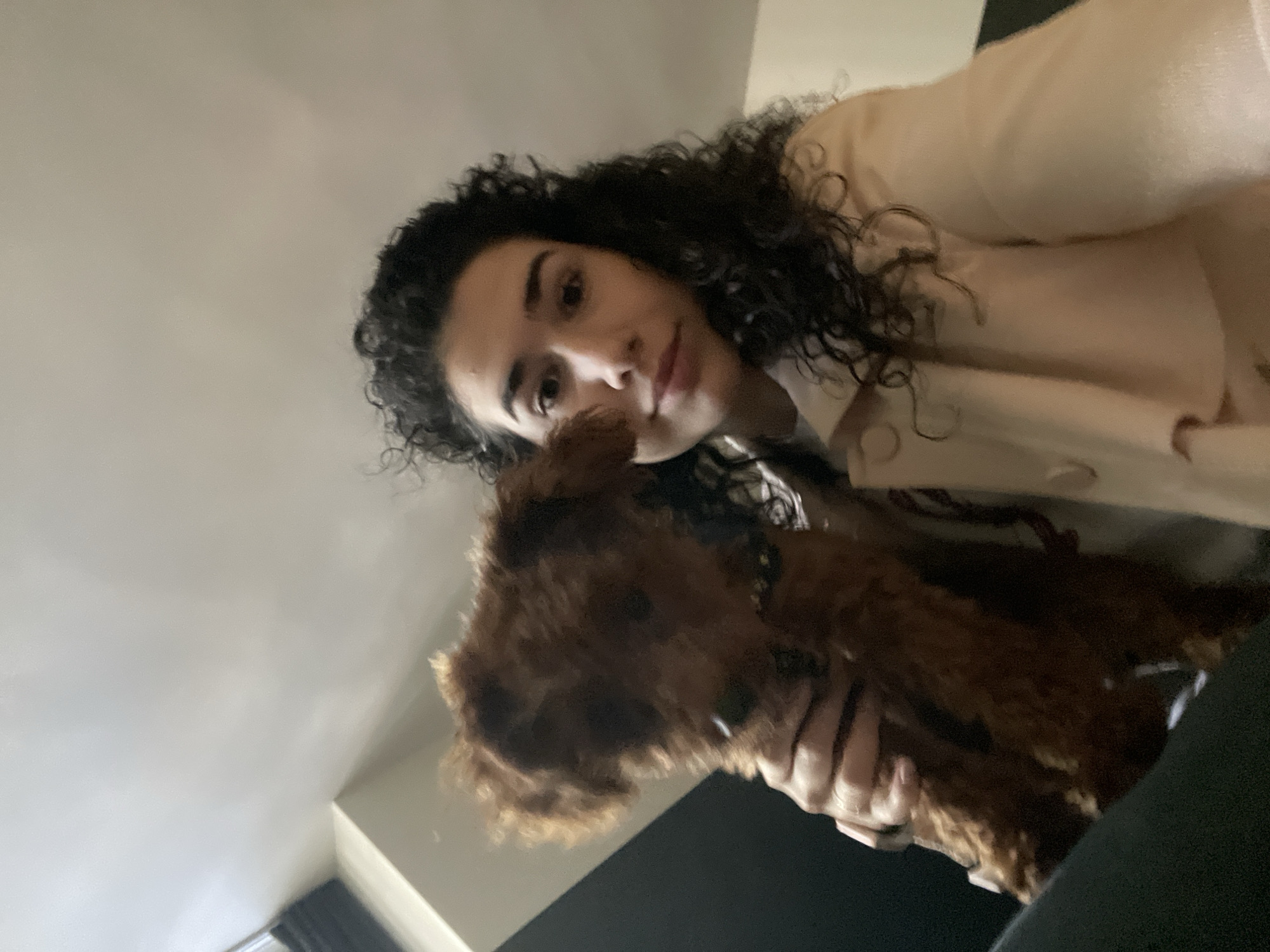 A person with curly dark hair and light skin is holding a brown, curly-haired dog. The person is wearing a light-colored jacket. The background consists of a white wall. The location cannot be identified within Sofia, Bulgaria based on this image.