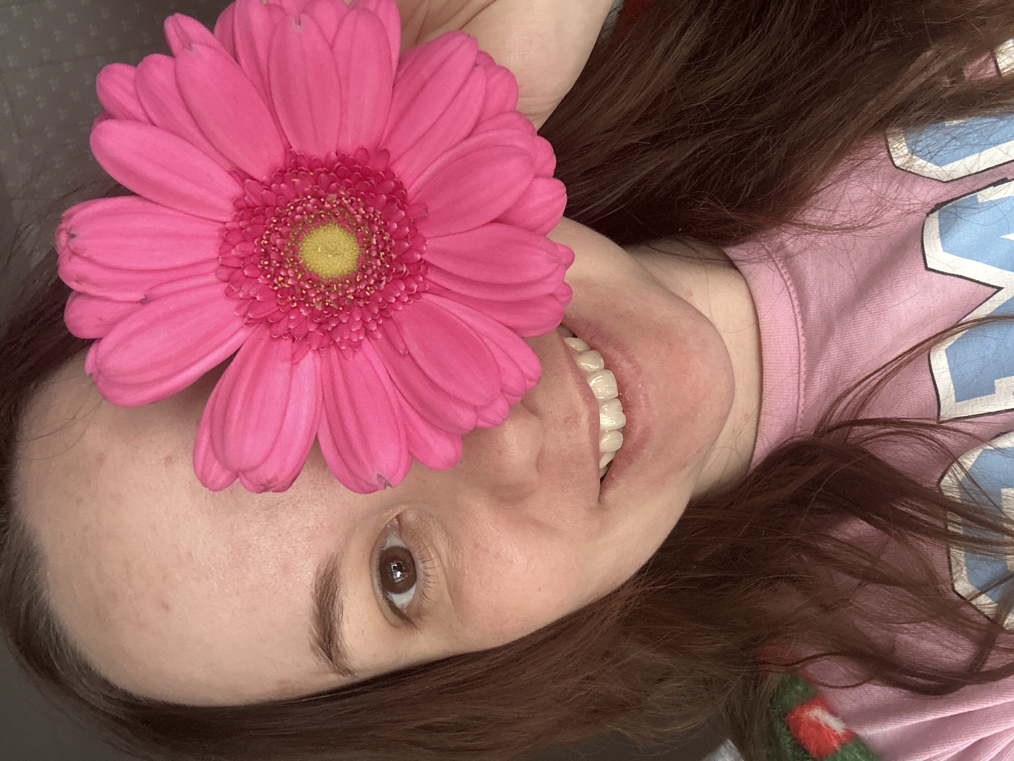 A woman is lying on her back, facing upward. She is holding a large, pink Gerbera daisy above her forehead. The flower has a bright yellow center. The woman has brown hair and fair skin. She is wearing a light pink t-shirt with blue lettering. She is smiling, revealing her teeth. The background is indistinct. The location is Unknown, Unknown.