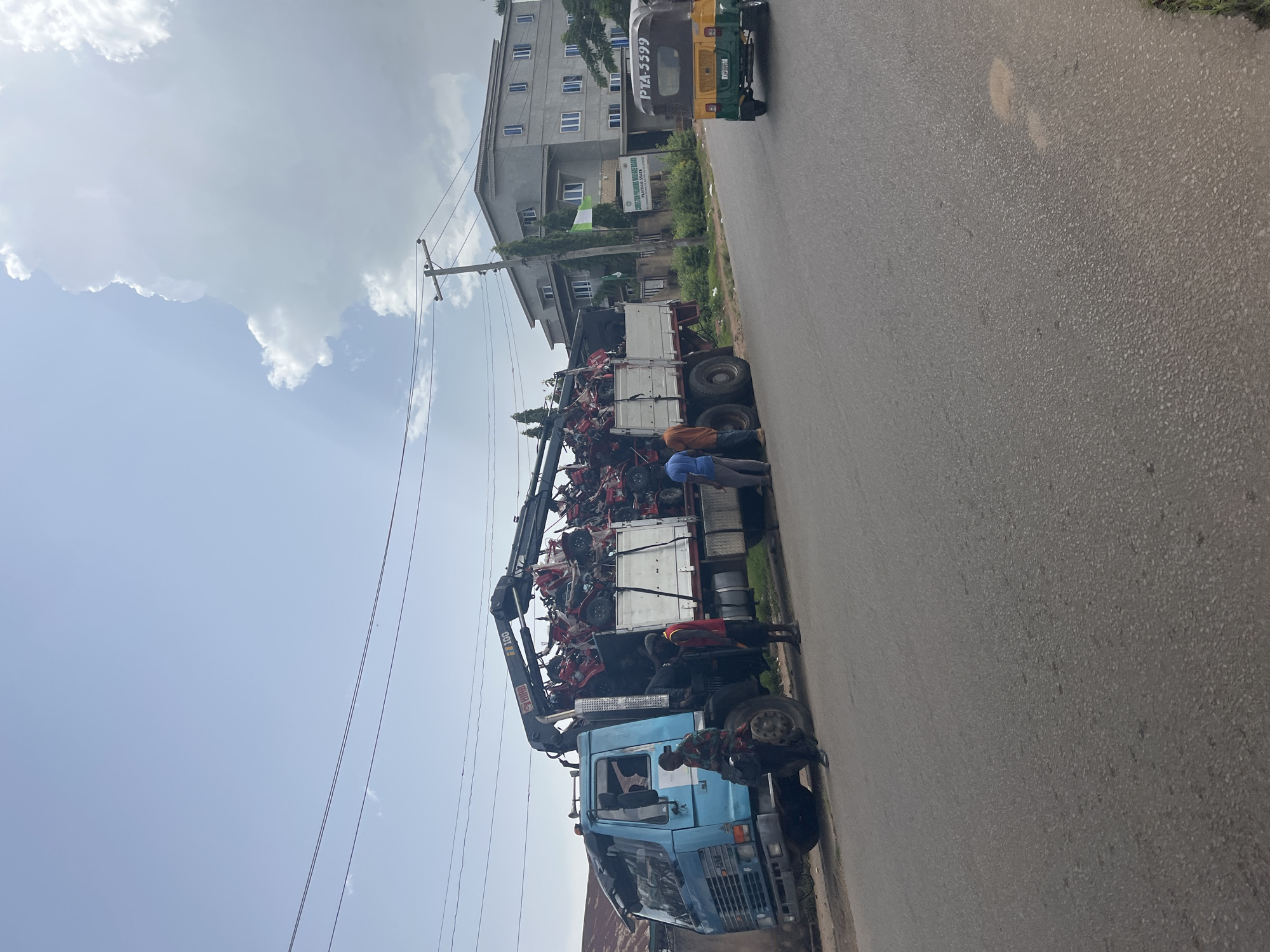The image captures an outdoor scene on a sunny day, likely in Jos, Nigeria. The main subjects are a blue flatbed truck with a crane attachment, laden with several red and white agricultural machines, possibly tillers or similar farming equipment. Several individuals are visible around the truck, appearing to be involved in loading or unloading the machinery. One person in an orange shirt is standing near the back of the truck, while others in blue shirts are situated on the ground or near the machinery.
The setting is a paved road, with a large, multi-story building in the background. The building appears to be a commercial or residential structure, with numerous windows. Adjacent to the building is a sign board that reads "SOLID MINERALS DEVELOPMENT BOARD" and below it, "GURARA COMPANY LIMITED". A Nigerian flag is also visible, attached to the building. In the distance, a yellow and green tuk-tuk with license plate "PTA-5599" is driving away. Overhead, power lines are strung across the sky, and there are scattered white clouds against a bright blue sky. The overall impression is one of a busy, operational day, likely related to the distribution or transportation of agricultural equipment in a developed area.