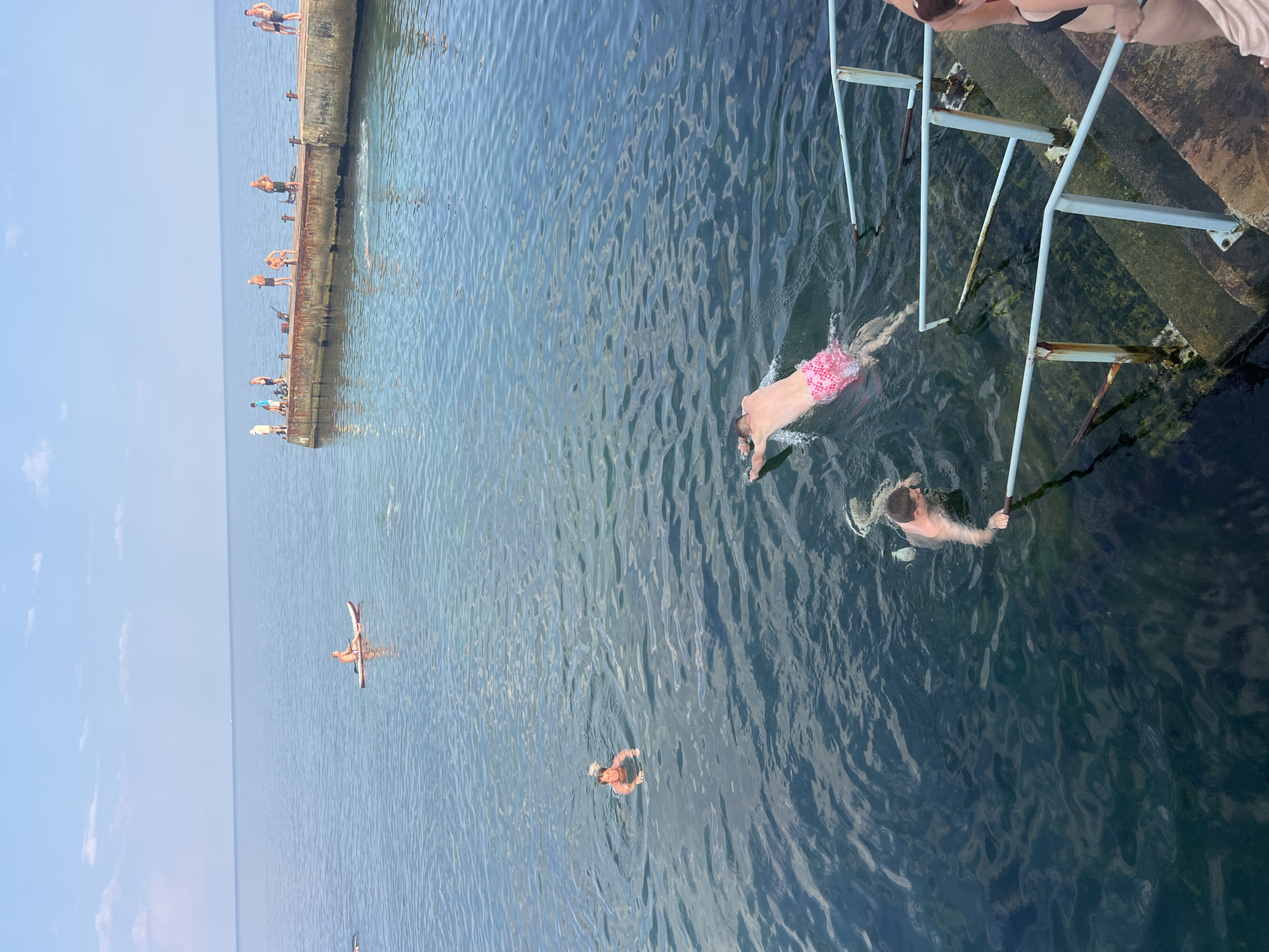 In Odesa, Ukraine, on a sunny day, several people are enjoying the water. The scene depicts a concrete pier extending into the calm, clear sea. On the pier, a group of individuals are standing, some appearing to be fishing. In the water, a person is paddleboarding, and several others are swimming. One man is swimming with pink patterned shorts, and another is visible near a set of concrete steps with railings leading into the water. The sky is blue with a few scattered clouds, and the horizon is visible in the distance.