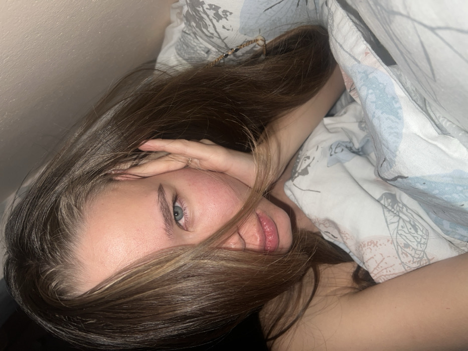 A young woman with light skin and blue eyes lies in bed, partially covered by a patterned duvet. Her brown hair with blonde highlights cascades around her face. One hand rests on her forehead, partly obscuring her features. She looks directly at the camera. Her lips are full and slightly parted. The lighting is soft, creating a cozy, intimate atmosphere. The image is rotated to the left slightly.