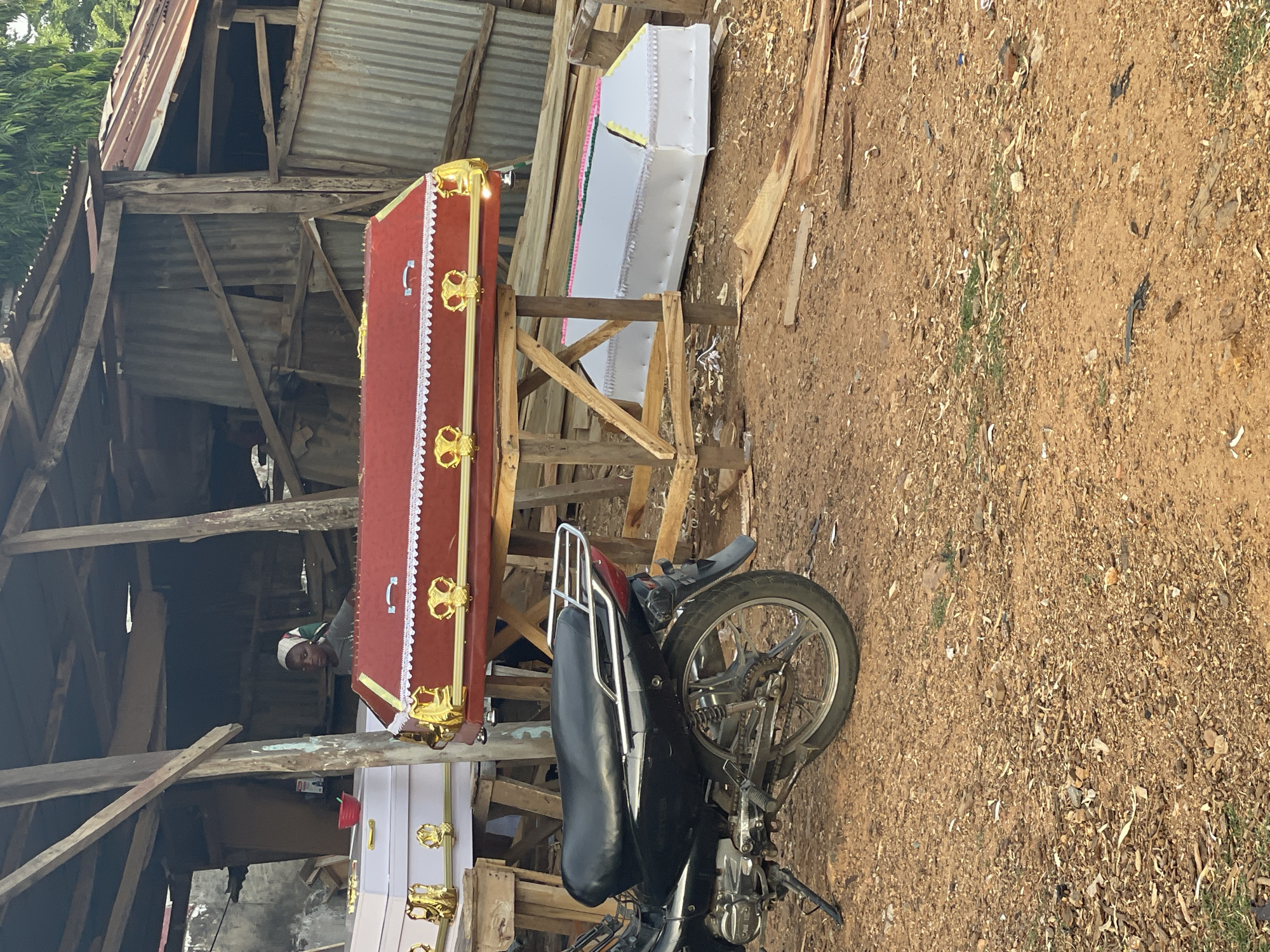 The scene is likely in Akwanga, Nigeria. The image depicts a dirt yard with wooden structures, possibly workshops or sheds, constructed from rough-hewn lumber and corrugated metal. Two coffins are elevated on wooden stands; one is red with gold-colored decorative hardware and white trim, and the other is white with similar decorative features. A black motorcycle is parked in front of the stands and partially obscures the view of the coffins. A person with a white head covering is standing behind the red coffin, peeking through the wooden structure. The ground is covered in loose dirt and wood debris.
