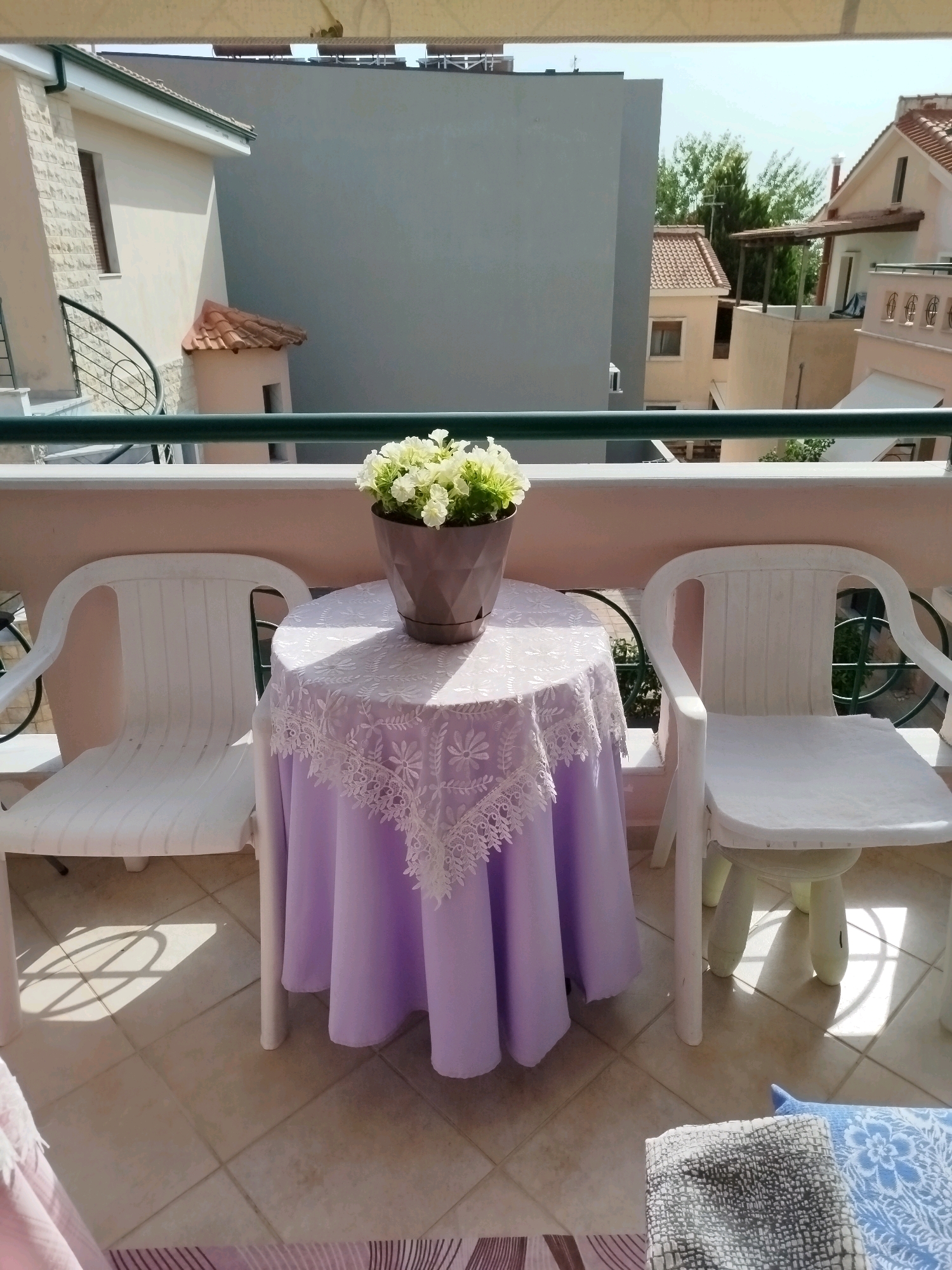 This is a medium shot, taken from a balcony overlooking a residential area in Sithonia Municipal Unit, Greece, on a sunny day. The foreground features a round table draped with a pale purple tablecloth adorned with white lace trim. A potted plant with white flowers sits in the center of the table. Two white plastic chairs, one on each side of the table, are also visible. The balcony floor is tiled. In the background, several buildings with terracotta roofs and cream-colored walls are visible, along with lush green trees and a clear blue sky. The scene suggests a peaceful outdoor seating area.