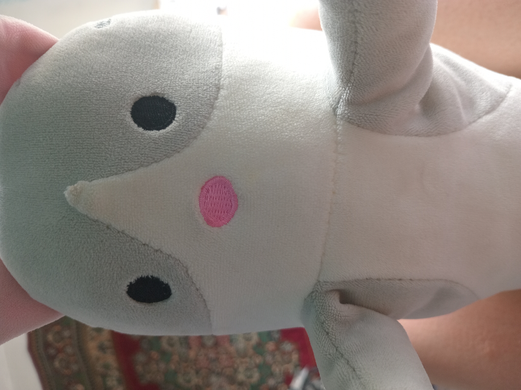 A plush toy animal, likely a cat or mouse, is shown in a close-up, head-on shot. The toy is predominantly light gray and white, with pink ears. It features black oval eyes and a pink circular nose embroidered onto its face. The toy's arms are visible, one on each side, extending outwards slightly. The background is blurred and appears to be an indoor setting with a patterned rug partially visible on the left and an area of light, possibly a window, on the right. The lower portion of the image shows skin, suggesting the toy is being held by a person.
