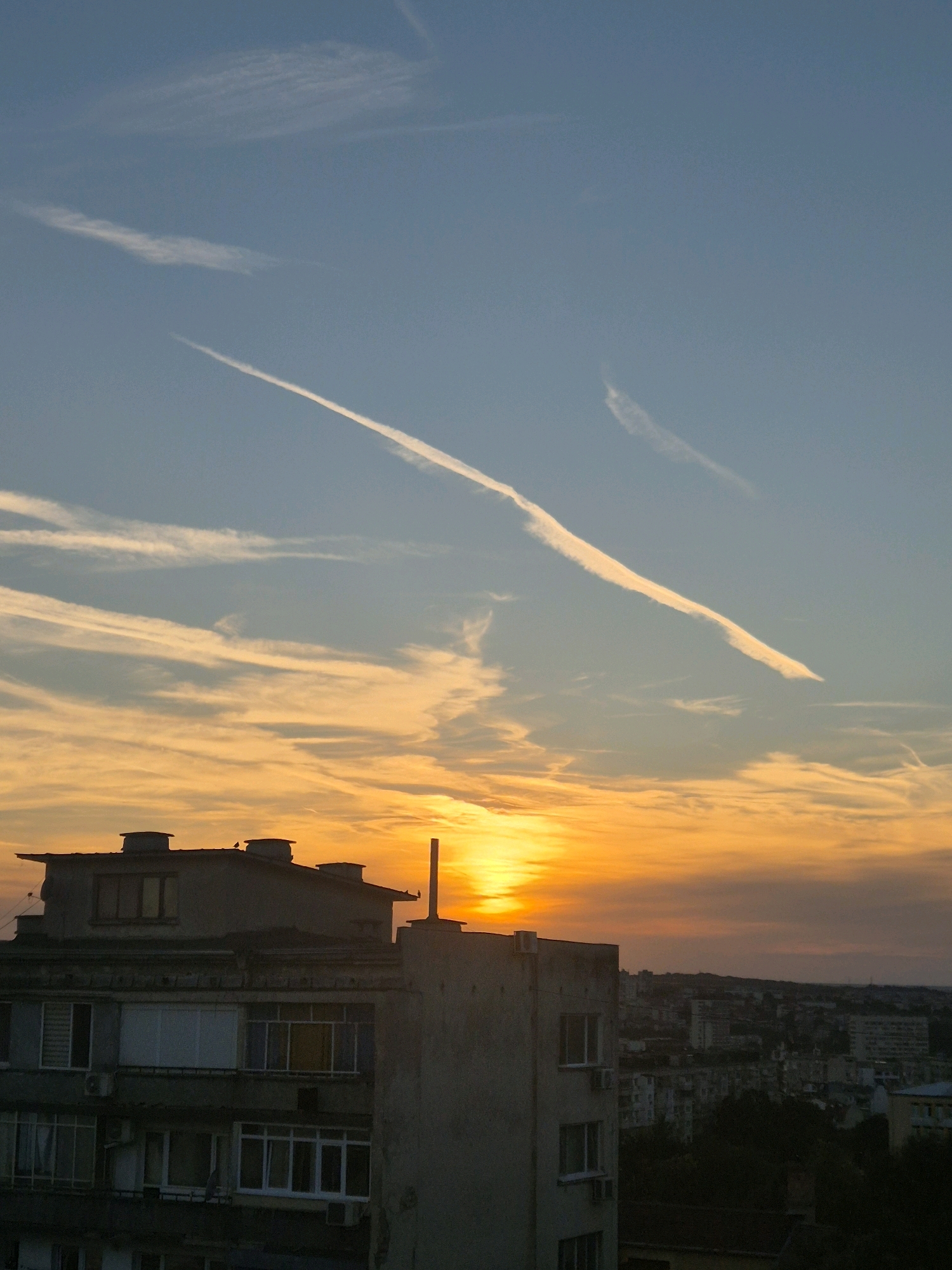 The image depicts a city skyline at sunset. The sky is a gradient of blue, orange, and yellow, with streaks of white clouds. The sun is visible near the horizon, casting a warm glow. In the foreground, a multi-story building with a flat roof and several windows is silhouetted against the sky. The building appears to be made of concrete. There are also two smaller buildings or structures on the roof of the main building. In the background, the city extends into the distance, with various buildings and trees visible. The overall impression is of an urban environment at the end of the day.