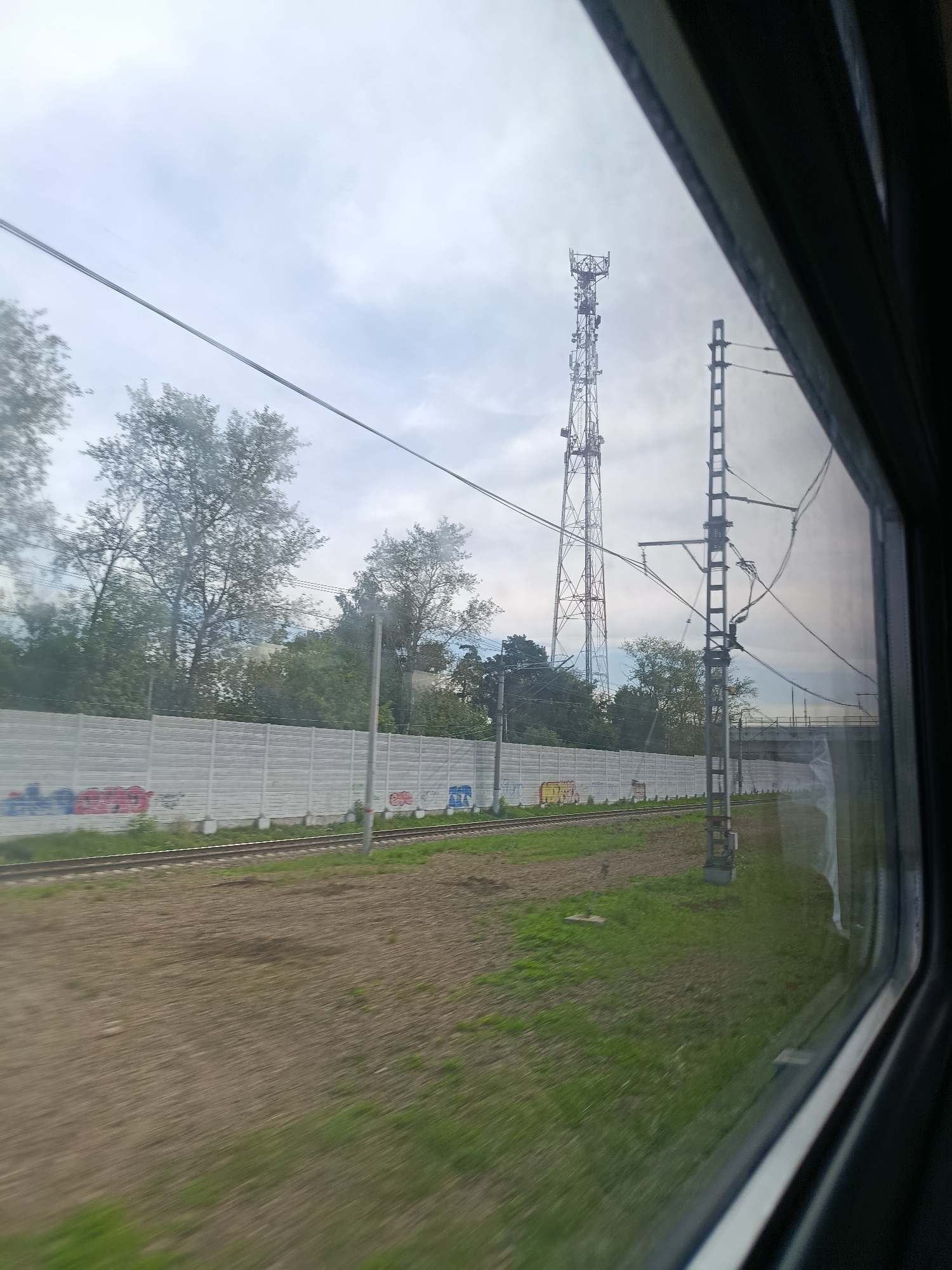 The image captures a view from a train window in Balashikha, Russia. A tall, lattice telecommunications tower is visible in the distance, silhouetted against a cloudy sky. In the foreground, railroad tracks run alongside a white sound barrier wall adorned with graffiti. The ground beside the tracks is covered in patchy grass and dirt, showing motion blur indicating the train is in transit. A utility pole with overhead wires is also present near the tracks. Trees are scattered throughout the landscape, and a bridge or overpass is dimly visible in the far right of the frame. The window frame of the train is visible on the right side of the image, creating a vignette effect.