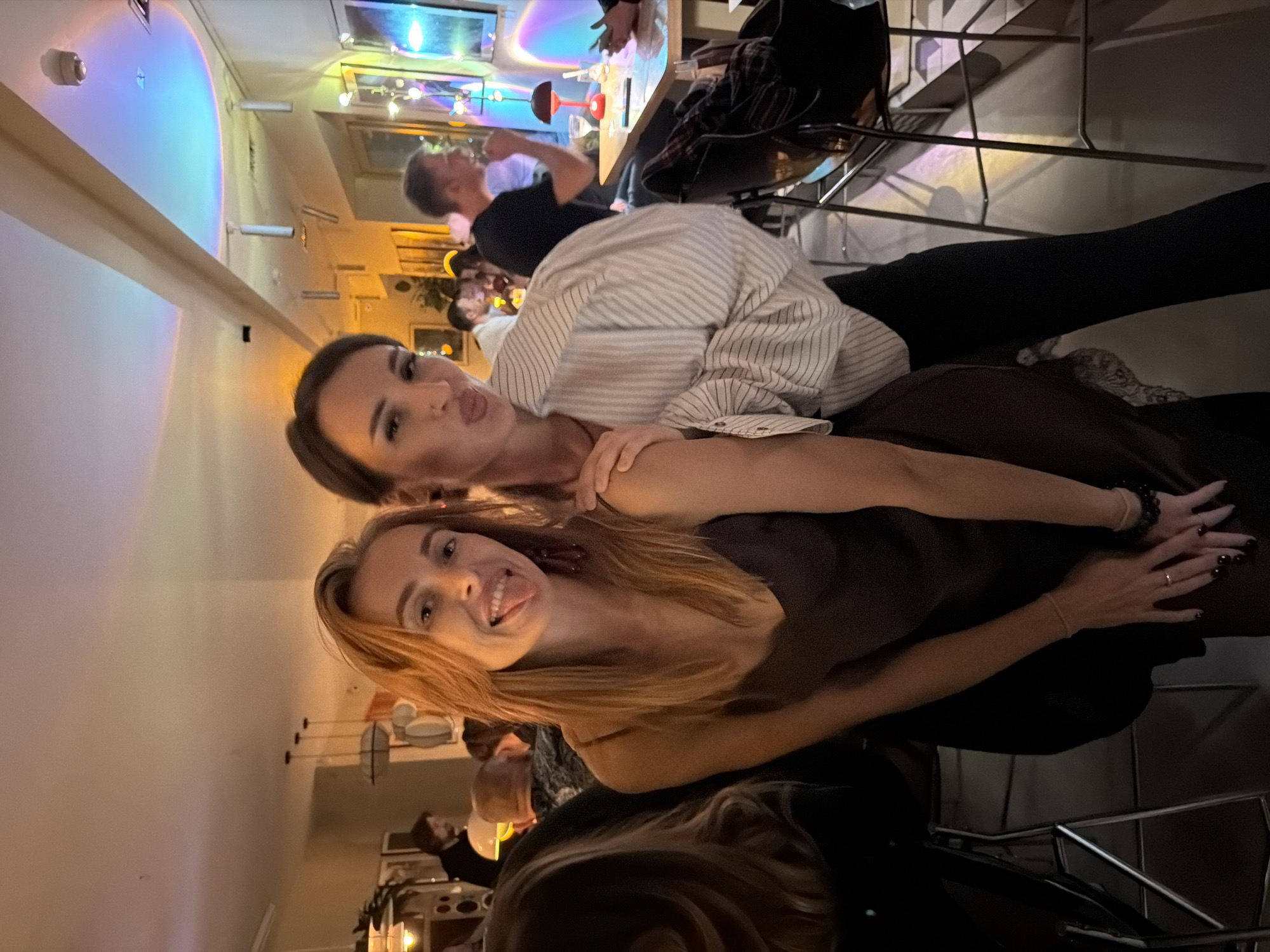 The scene is an indoor social gathering, likely in a restaurant or bar in Krasnoyarsk, Russia. Two women are prominently featured, posing for the camera and sticking out their tongues in a playful manner. One woman has light brown hair and wears a dark brown dress. The other woman has dark hair pulled back and wears a striped shirt. They are seated, possibly on bar stools. In the background, other people are present, engaged in conversations and activities related to dining and socializing. The lighting is mixed, with some areas brightly lit and others darker. A bar or countertop is visible, suggesting a commercial establishment.