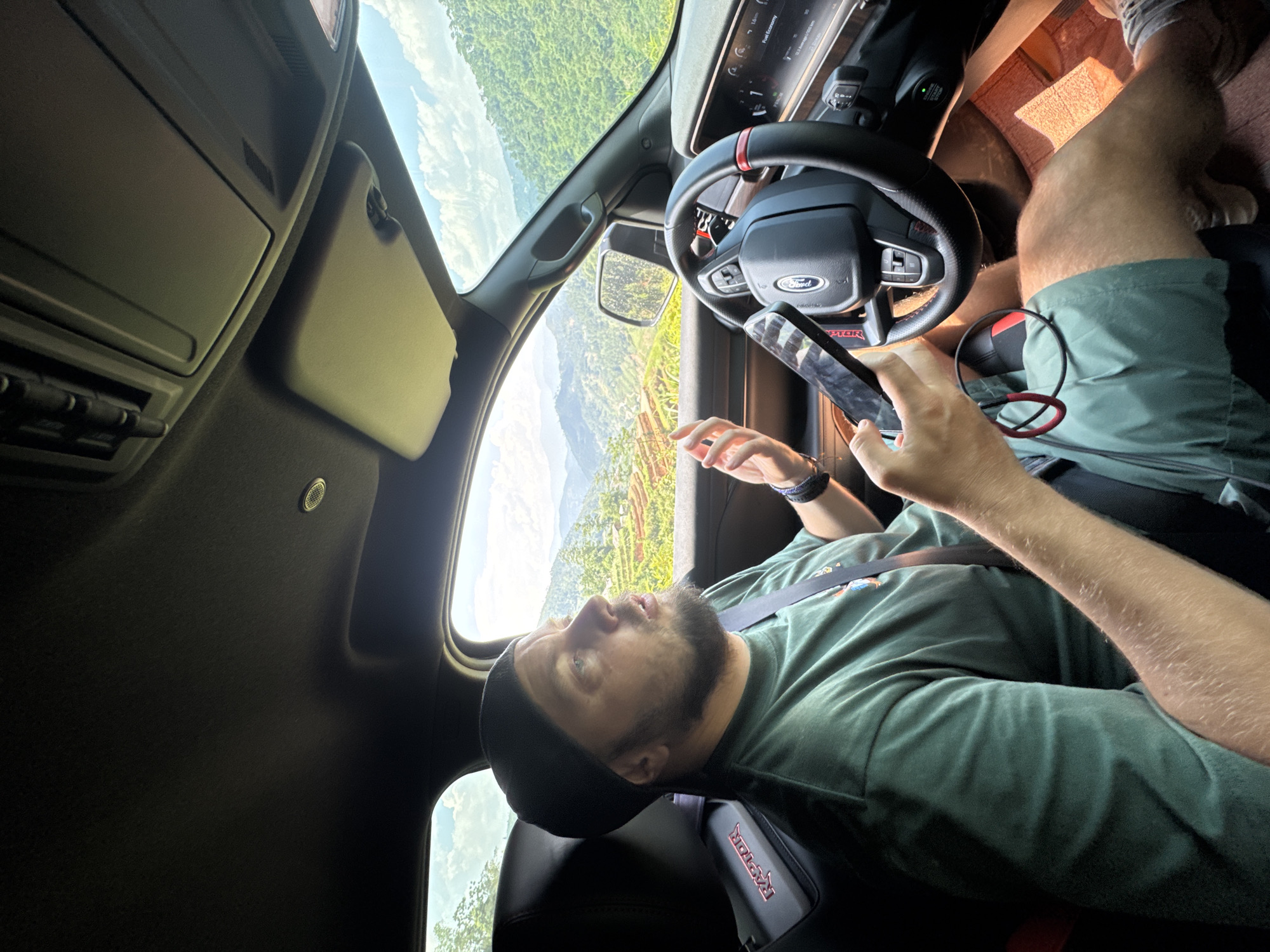 A man is sitting in the driver's seat of a Ford pickup truck. He is wearing a dark beanie, a green T-shirt, and green shorts. He is holding a smartphone in his hand and appears to be looking at it. The truck's interior is black. The steering wheel has a Ford logo in the center. Through the windshield, a landscape of hills and mountains can be seen, possibly in Vinh Quang, Vietnam.