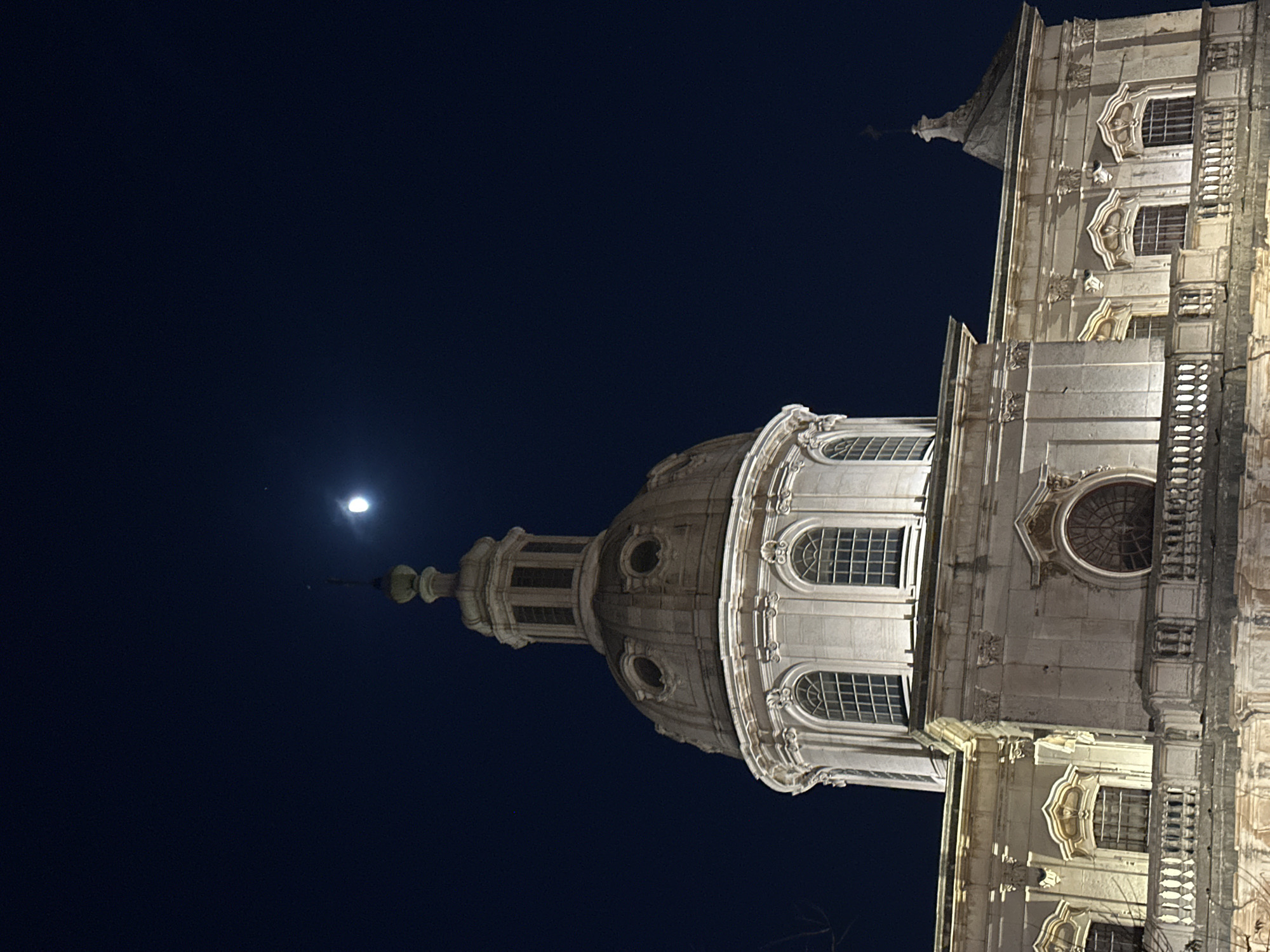This is a vertical, nighttime photograph of the dome and facade of a large, ornate building, likely a church or basilica. The building is made of light-colored stone and features intricate architectural details such as arched windows, columns, and decorative molding. The dome is prominent, with multiple levels and windows, and is topped by a spire. A balcony with a railing runs along a portion of the facade.
The sky is a deep, dark blue, indicating nighttime. A bright, full moon is visible in the upper left quadrant of the image, casting a soft glow. A small, faint light source, possibly a star or a distant light, is also visible near the moon. The building appears to be illuminated by artificial lighting, highlighting its architectural features against the night sky.
The overall mood is serene and majestic, emphasizing the grandeur of the structure. There are no people visible in the image. The setting is urban, with the architecture suggesting a historical European city. Given the location context of Lisbon, Portugal, this could be a famous landmark such as the Monastery of São Vicente de Fora. No text is visible in the image.