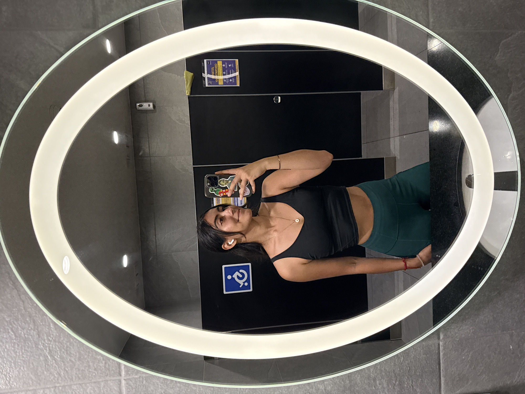 A person is visible in a reflection in an oval-shaped mirror with a ring light around it. The person is a woman with dark hair pulled back, wearing a black tank top and green leggings. She is holding a phone with a decorated case, presumably taking a selfie. She is wearing a necklace and bracelets. The background behind the woman in the mirror appears to be a bathroom with gray tile walls and black stall doors. There are signs on the stall doors, including one with a wheelchair accessibility symbol. There is also a small security camera on the wall. The mirror is presumably in a commercial or public restroom. There are no distinct landmarks visible to definitively determine the location in Puebla, Mexico.