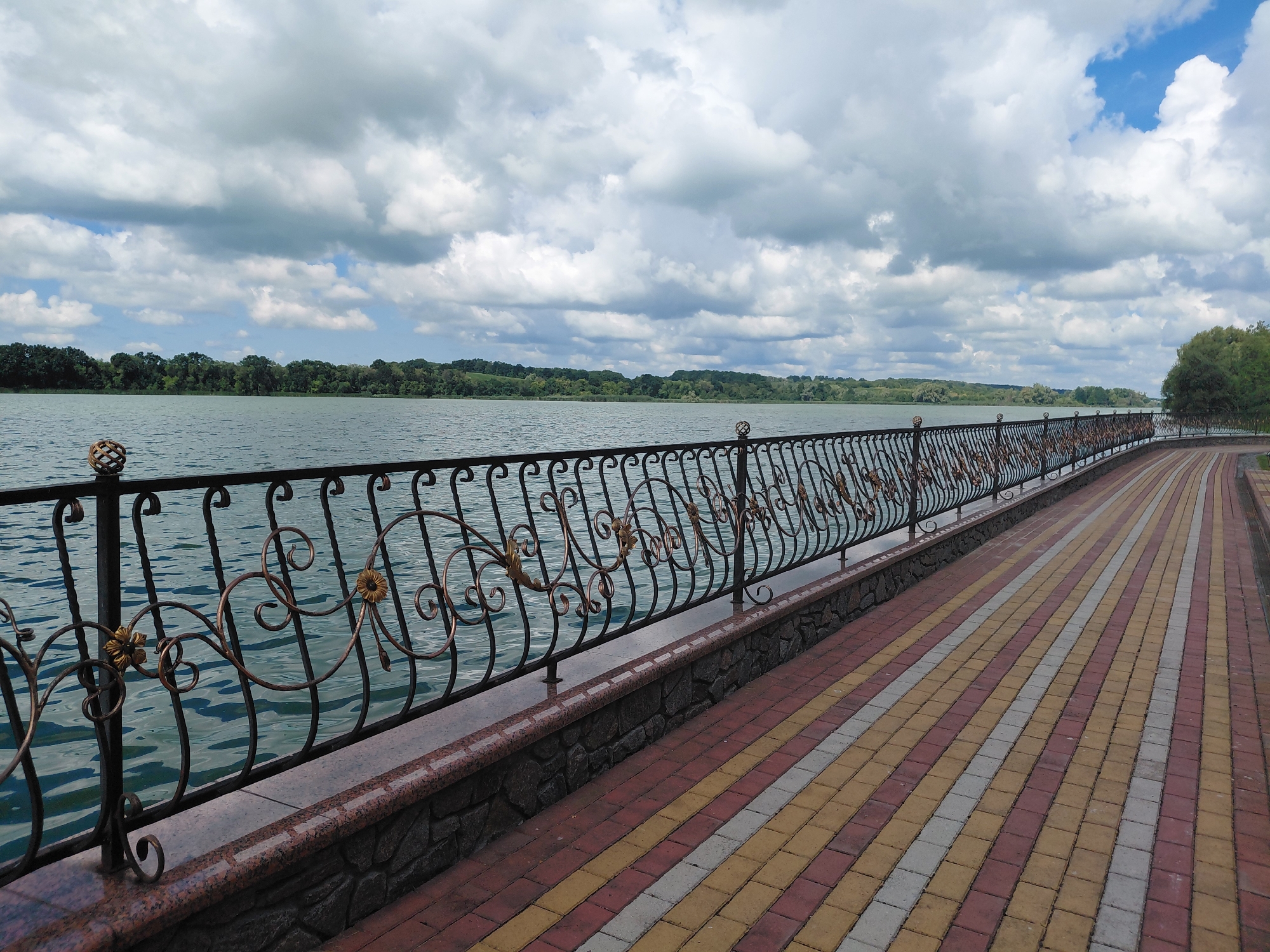 The image shows a waterfront promenade in Kovalivka, Ukraine, under a cloudy sky. A decorative black metal fence with ornate gold accents separates the promenade from a calm body of water. The promenade itself is paved with interlocking bricks in a pattern of red, yellow, and white. In the background, a treeline with lush green foliage borders the water, and the sky is filled with dramatic, textured clouds. The scene conveys a sense of public space and natural beauty.