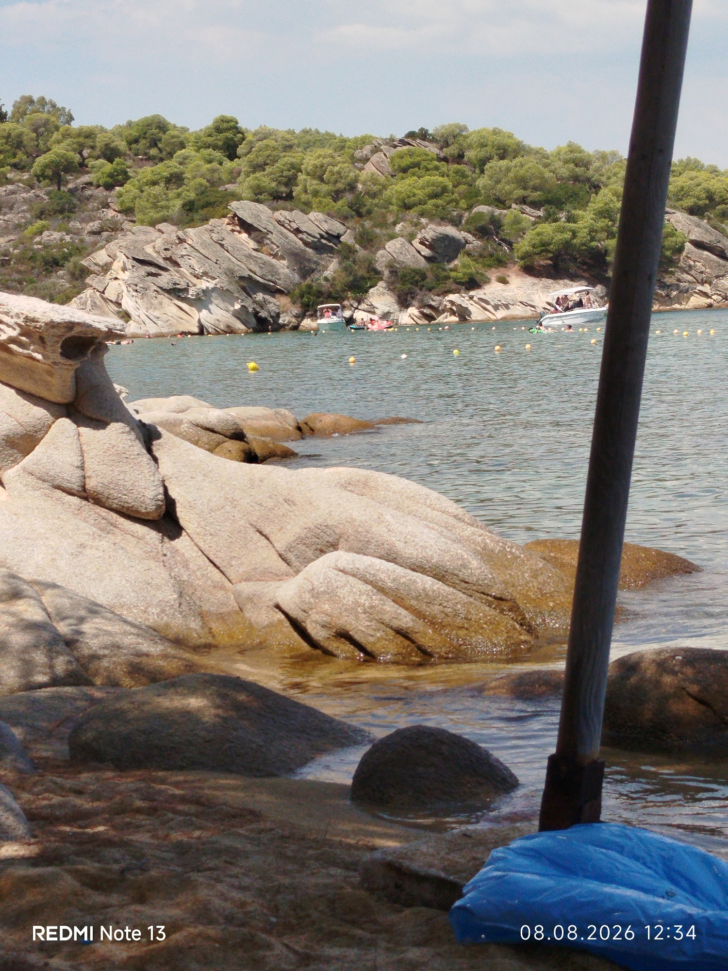 A photograph of a rocky beach in Sithonia Municipal Unit, Greece, on August 8, 2026, at 12:34 PM. The foreground features large, light-colored rocks with smooth, worn surfaces. Some of these rocks are partially submerged in clear, shallow water, revealing their texture and color. A dark, cylindrical object, possibly a wooden pole, is visible to the right, embedded in the sand and water. At the base of the pole, a bright blue, soft object, potentially a bag or cushion, rests on the shore.
In the middle ground, the water stretches out, dotted with numerous small, yellow buoys. A white boat with several people on board is visible in the distance, navigating the water. Beyond the water, the landscape rises to a hill covered with dense green trees and rugged, rocky formations. The sky above is a pale blue with scattered, wispy clouds. The overall scene depicts a sunny day at a coastal location, likely a popular spot for swimming and recreational activities.