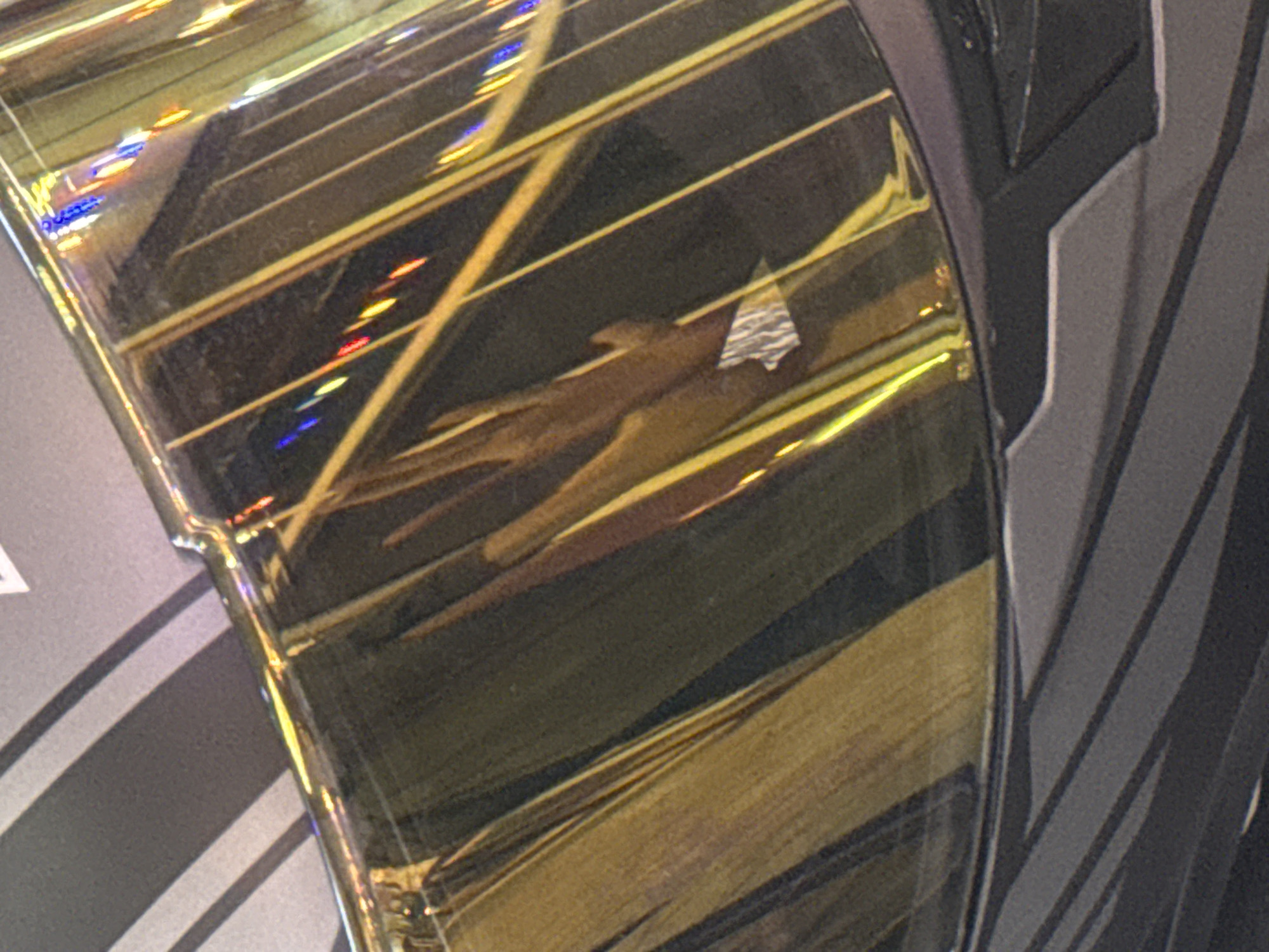 This is a close-up, angled, and partially blurred photograph of a reflective surface, likely part of a vehicle or decorative object. The surface has a metallic, gold-like sheen with prominent yellow and brown striations, possibly indicating the texture or pattern of the object. There are reflections of colorful lights, including red, blue, and yellow, suggesting an outdoor or brightly lit environment, possibly a street at night or an area with decorative lighting. In the foreground, the lower part of the image shows a matte gray surface with diagonal lines, which appears to be a contrasting material or a different section of the same object. The image is focused on the reflective and textured elements, with the background and specific context of the location being obscured by the close-up perspective and reflections. There are no discernible people, animals, or specific landmarks visible in this crop, making it impossible to ascertain its location within Karon, Thailand, or any other specific geographical area.