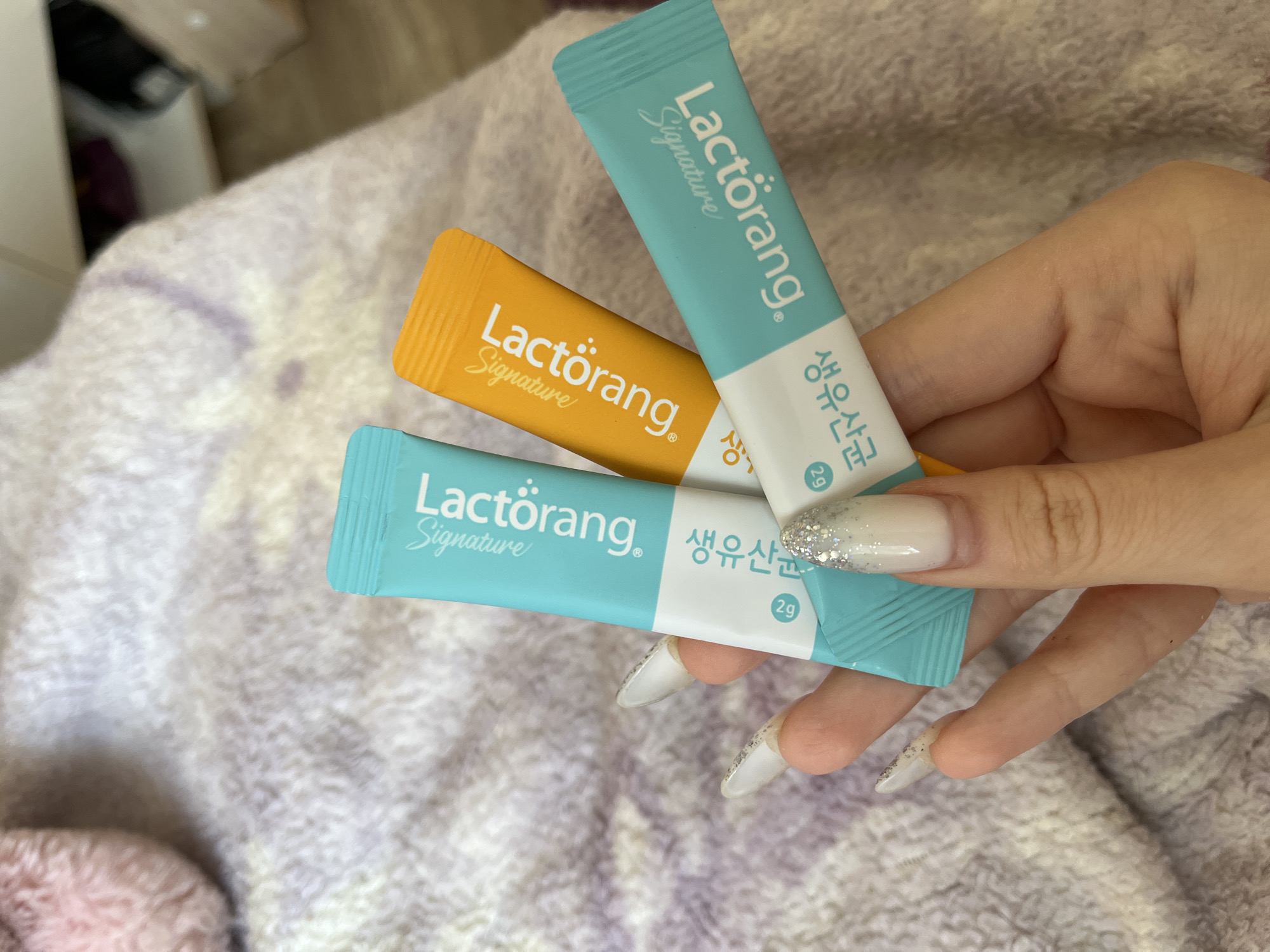 A close-up, eye-level shot shows a person's hand holding three small, individually wrapped packets of a supplement. The packets are turquoise, orange, and turquoise. The turquoise packets have white text that reads "Lactorang Signature" and Korean text below it. The orange packet has similar white text. The hand has long, artificial nails with a glittery, French manicure design. The background is a soft, textured, light-colored blanket with some purple hues. The image is well-lit, with the focus on the hand and the packets. This image was taken in a domestic setting and provides no information about the city of Ivanovo, Russia.