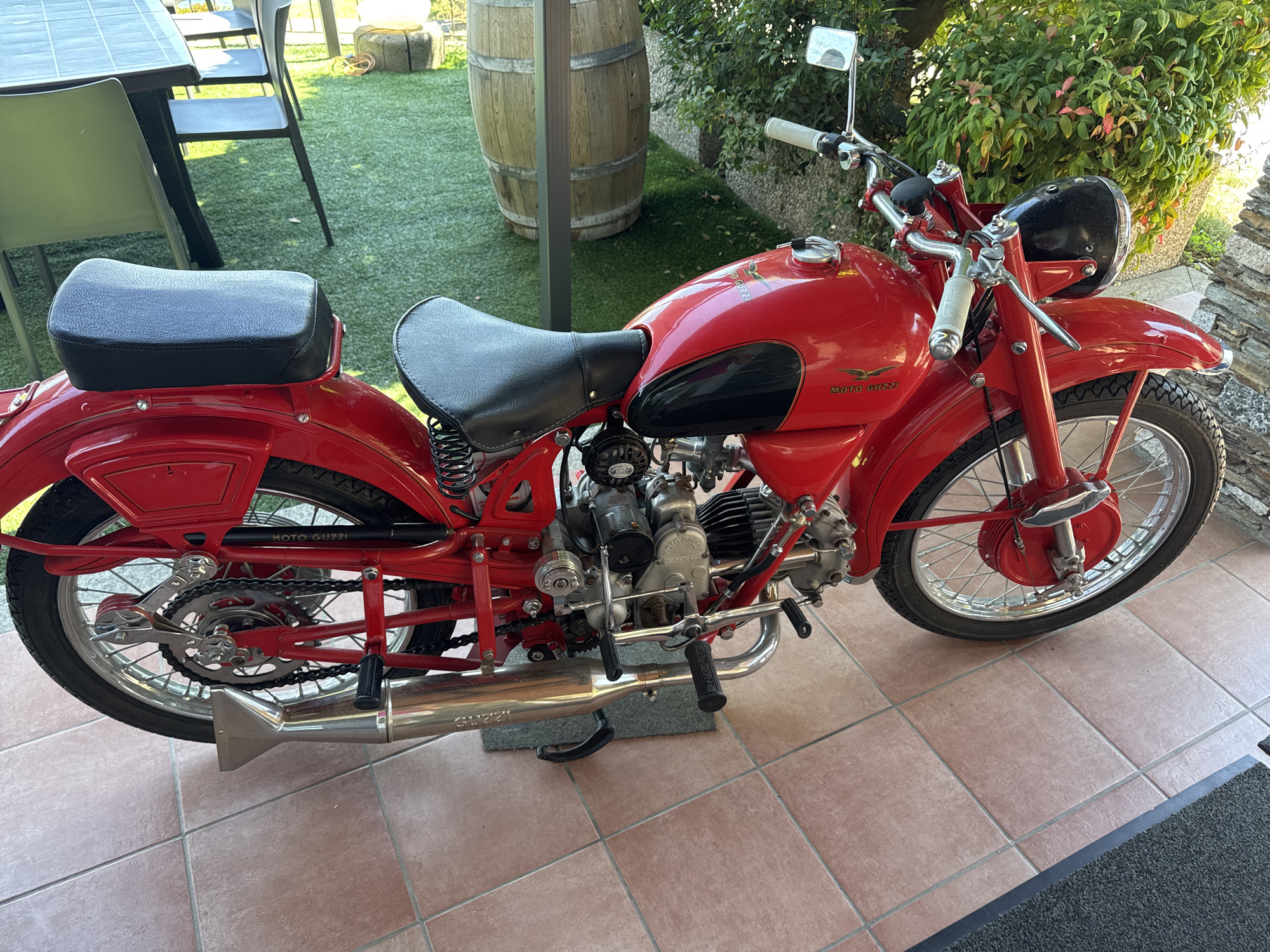 A vintage red Moto Guzzi motorcycle is parked on a tiled patio. The motorcycle is in good condition with a black leather seat and chrome accents. In the background, there is a large wooden barrel, a green artificial lawn with tables and chairs, and some bushes. The scene appears to be outdoors, possibly in a courtyard or garden area. The location is identified as Carciano, Italy. There are no people or animals visible in the image.