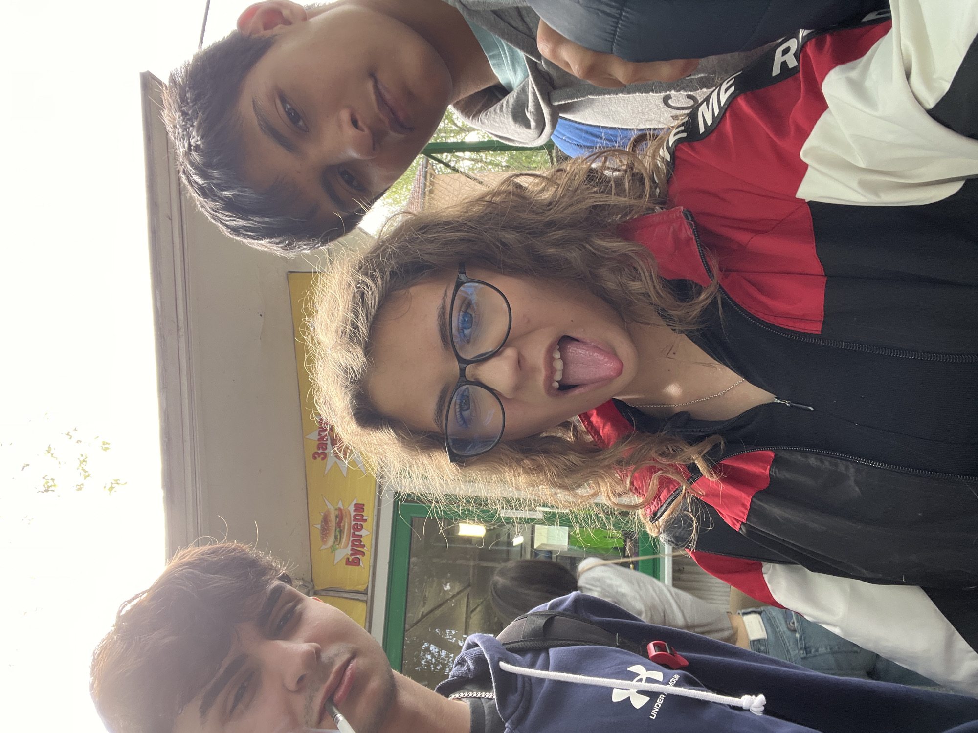A low-angle selfie captures three young individuals in what appears to be an outdoor setting in Plovdiv, Bulgaria, during the daytime. The image is rotated 90 degrees counter-clockwise.
In the upper portion of the frame (left after rotation), a young male with short dark hair looks towards the camera with a neutral expression. He is wearing a light-colored, possibly grey or light blue, hooded sweatshirt.
Dominating the center is a young female with long, wavy, light brown hair and black-rimmed glasses, revealing blue eyes. She playfully sticks out her tongue and looks directly at the camera. She is wearing a distinctive jacket with a black, red, and white color-block design. Part of a word, "REME" or "PREME," is visible on a black band on her jacket.
In the lower part of the frame (right after rotation), another young male with dark hair and some facial stubble is visible. He holds a white object, possibly a vape pen or cigarette, near his mouth. He is wearing a dark blue hoodie with "UNDER ARMOUR" text and its logo clearly visible on the sleeve.
The background shows a brightly lit area suggesting sunlight, along with blurred figures of other people, indicating a public space. A prominent yellow sign is visible behind the subjects, featuring red Cyrillic text that reads "Закуски" (Snacks) and "Бургери" (Burgers), accompanied by a graphic of a hamburger. This suggests the setting is near a food stall or kiosk.