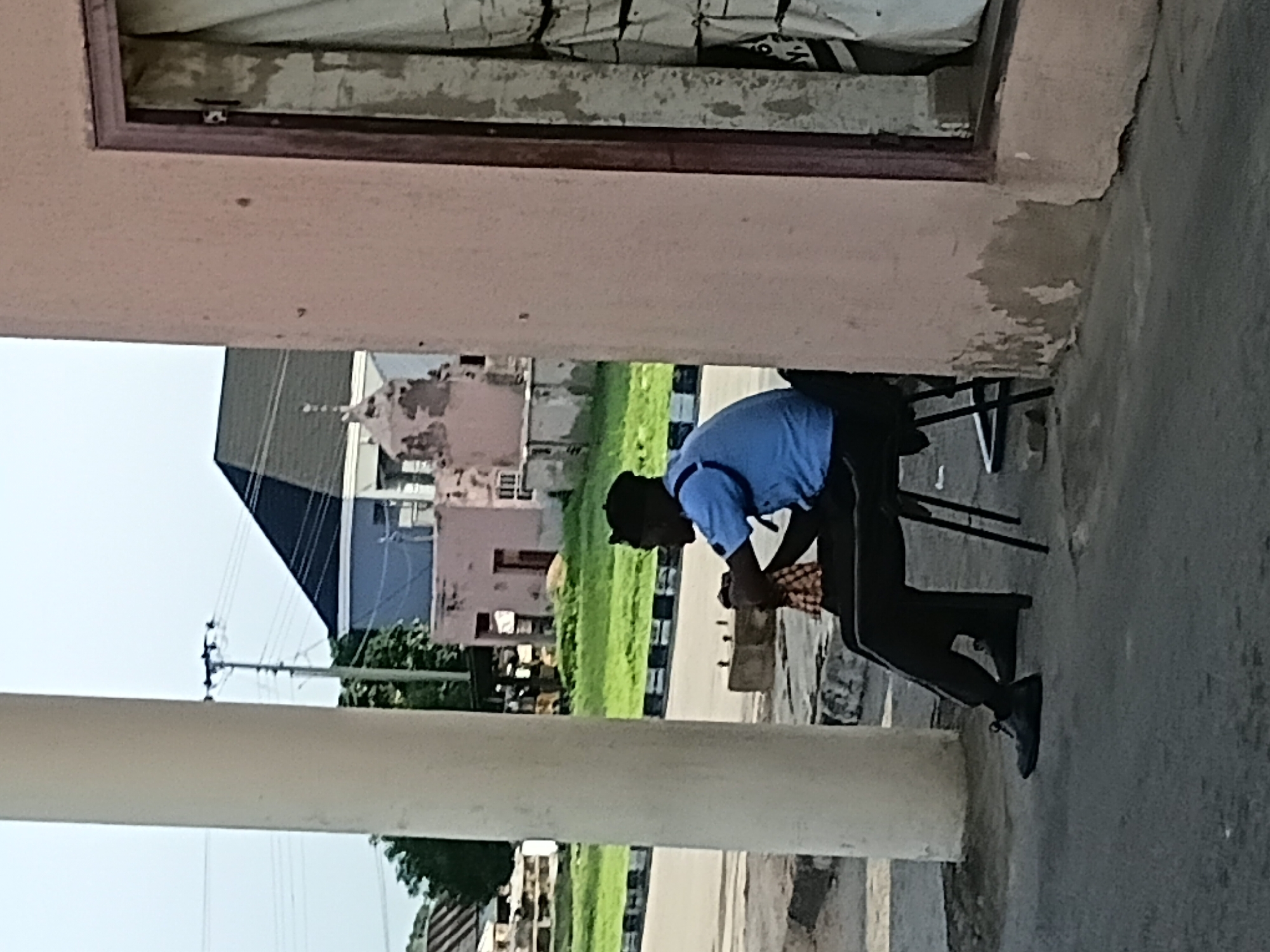 A man wearing a blue short-sleeved shirt, dark trousers, and a black cap is sitting on a black plastic chair outdoors. He is holding a checkered cloth in his lap with his left hand and his right hand is resting on his lap. He is facing to the right, in profile. The man is seated next to a large, light-colored cylindrical pillar. In the background, there is a road with black and white painted borders. Beyond the road, there is a grassy area with some trees. Further in the background, there are buildings, including a pink building with a dark roof, and a lighter-colored building with a small domed structure on top. Electrical poles and wires are visible in the sky. The foreground shows a grey paved surface. The image appears to be taken in Jalingo, Nigeria.