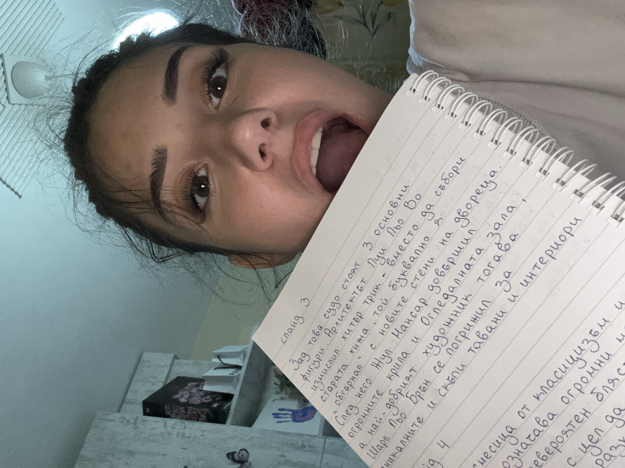 The image shows a young woman with her mouth open, holding a notebook with handwritten text. The notebook is in Bulgarian and appears to be a school assignment or notes. The woman is indoors, with a white ceiling and a wall with some artwork in the background. The woman's face is the primary focus, with her eyes looking directly at the camera. The notebook is positioned in front of her, obscuring part of her face. The overall lighting suggests an indoor environment with artificial light. The text in the notebook appears to be a narrative passage, possibly from a book or a story. The woman's expression suggests surprise or excitement. The text on the notebook includes words like "слайд 3", " архитект", "ръка", "рисунки", and "двореца", indicating that it might be related to architecture or art. The woman is not located in the city of Plovdiv, Bulgaria, as there are no visual cues to suggest this location.