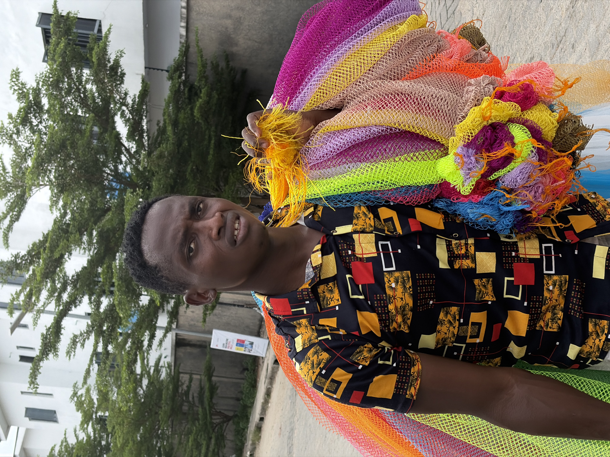 A man stands on a paved surface in Igboefon, Nigeria, holding a pile of colorful, net-like materials. The man is wearing a short-sleeved shirt with a patterned design in black, yellow, and red. The nets he is holding are in a spectrum of colors including pink, purple, yellow, orange, blue, green, and brown. Behind the man, there is a building with a tree in front of it. A sign is visible on the left.