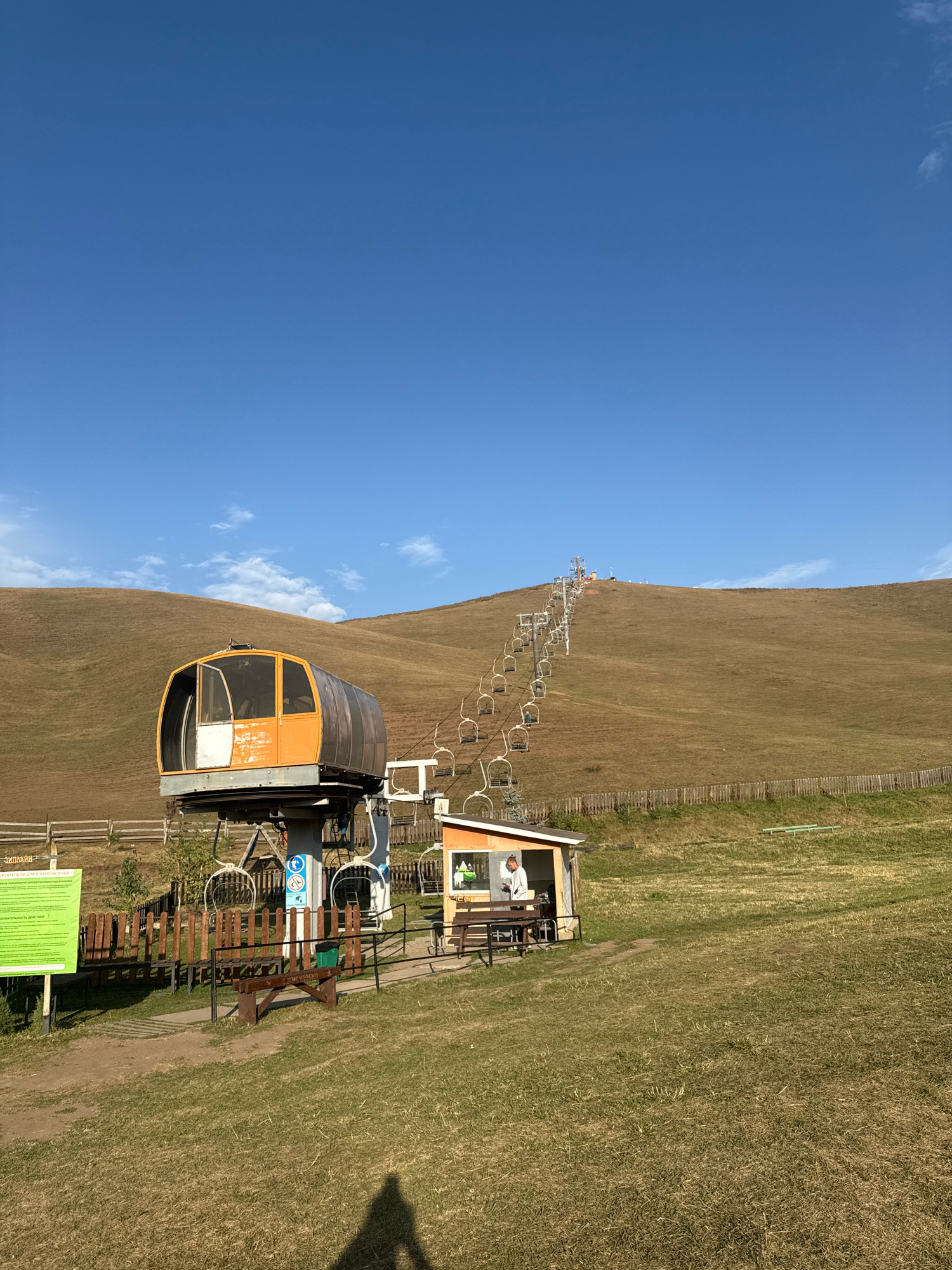 The image depicts a ski lift station on a grassy hillside under a clear blue sky with scattered clouds, in the location of Chunkurchak, Kyrgyzstan. The ski lift features multiple empty chairlifts ascending a gentle, brown-grass covered slope. In the foreground, an orange and gray ski lift station with a large, enclosed cabin is visible, positioned on a metal support. Adjacent to the station is a small, open-sided structure where a person in a white shirt is standing. There are wooden benches and a green trash bin near the ski lift. A wooden fence runs along the middle ground of the hillside. The overall environment suggests an outdoor recreational area in a mountainous or hilly region.