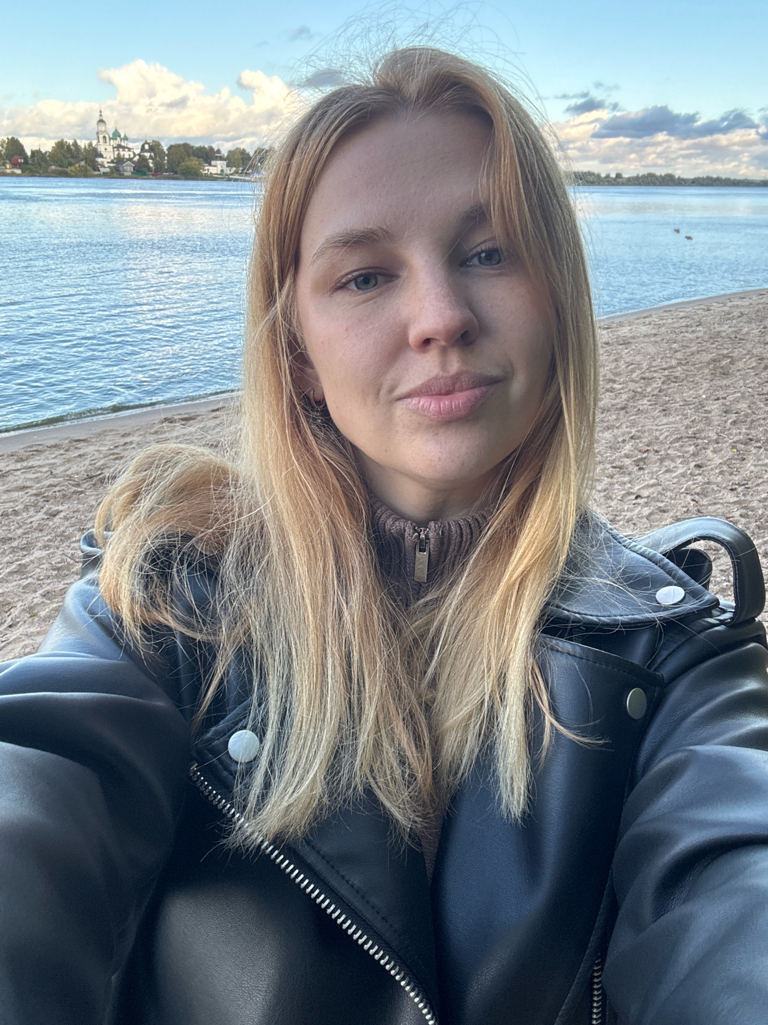 In this selfie taken outdoors, a young woman with long blonde hair is positioned in the foreground, facing the camera. She is wearing a black leather jacket over a brown turtleneck sweater. Her hair is falling loosely around her shoulders, with some strands obscuring parts of her face and clothing.
Behind her, a sandy beach extends to the edge of a body of water, likely a river or lake, with ripples indicating gentle movement. In the distance, across the water, is a picturesque scene with a white building adorned with green domes and crosses, reminiscent of a church or monastery. This architectural style suggests a location in Russia, and given the iconic church structure, it is highly probable that this image was captured in Yaroslavl, Russia, specifically on the banks of the Volga River, possibly near the St. John the Baptist Church in Tolchkovo or a similar historical complex. The sky above is partly cloudy with hints of blue peeking through, suggesting a pleasant day. The overall atmosphere is serene and picturesque.