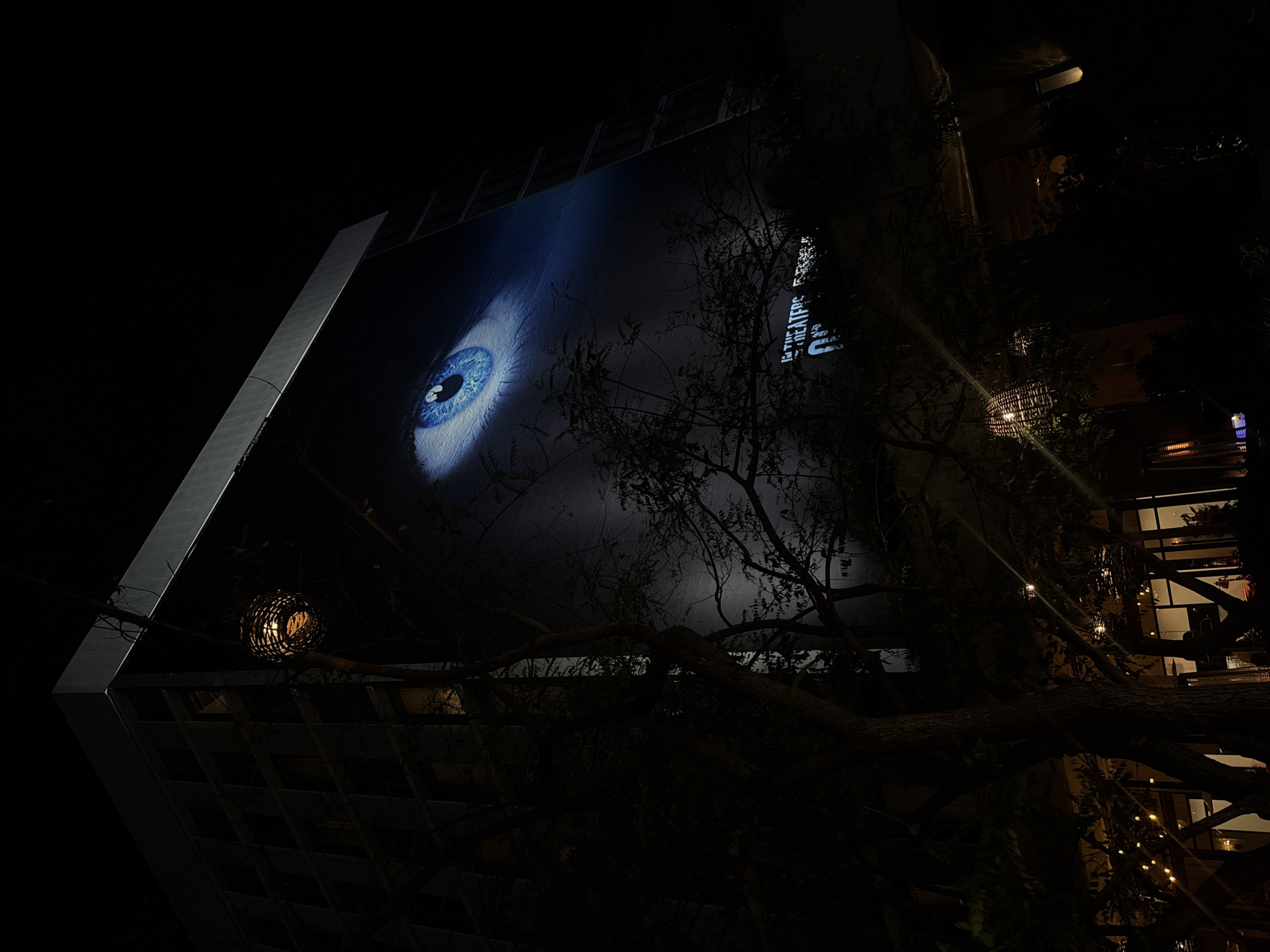 This media file presents a captivating nighttime scene in West Hollywood, United States of America. The primary subject is a colossal, intensely detailed blue eye projected onto the dark facade of a modern building, dominating the left and central portions of the frame. The eye features a bright blue iris, a prominent pupil, and surrounding white sclera, creating a striking focal point against the dark backdrop.
To the right of the eye, partially obscured by tree branches, is white text indicating a film promotion. The readable text clearly states "IN THE THEATERS SEPT 19" and "DON'T BREATHE 2", identifying the horror film being advertised.
The setting is dark, signifying nighttime, with the main light source emanating from the illuminated projection itself. Silhouettes of numerous tree branches and foliage crisscross the foreground and midground, partially obscuring parts of the building and the text, adding depth and a natural element to the urban environment. A distinctive spherical, caged light fixture hangs from a tree branch in the upper left section. Further to the right, through the dense foliage, glimpses of string lights and interior lighting from another building or establishment suggest a nearby social or commercial area.
The overall atmosphere is mysterious and intriguing, underscored by the intense gaze of the large eye and the deep shadows of the night. The building itself exhibits modern, geometric architectural elements, visible beneath and adjacent to the large projection. No specific human activity is visible, but the scene depicts an outdoor public advertisement in an urban context.