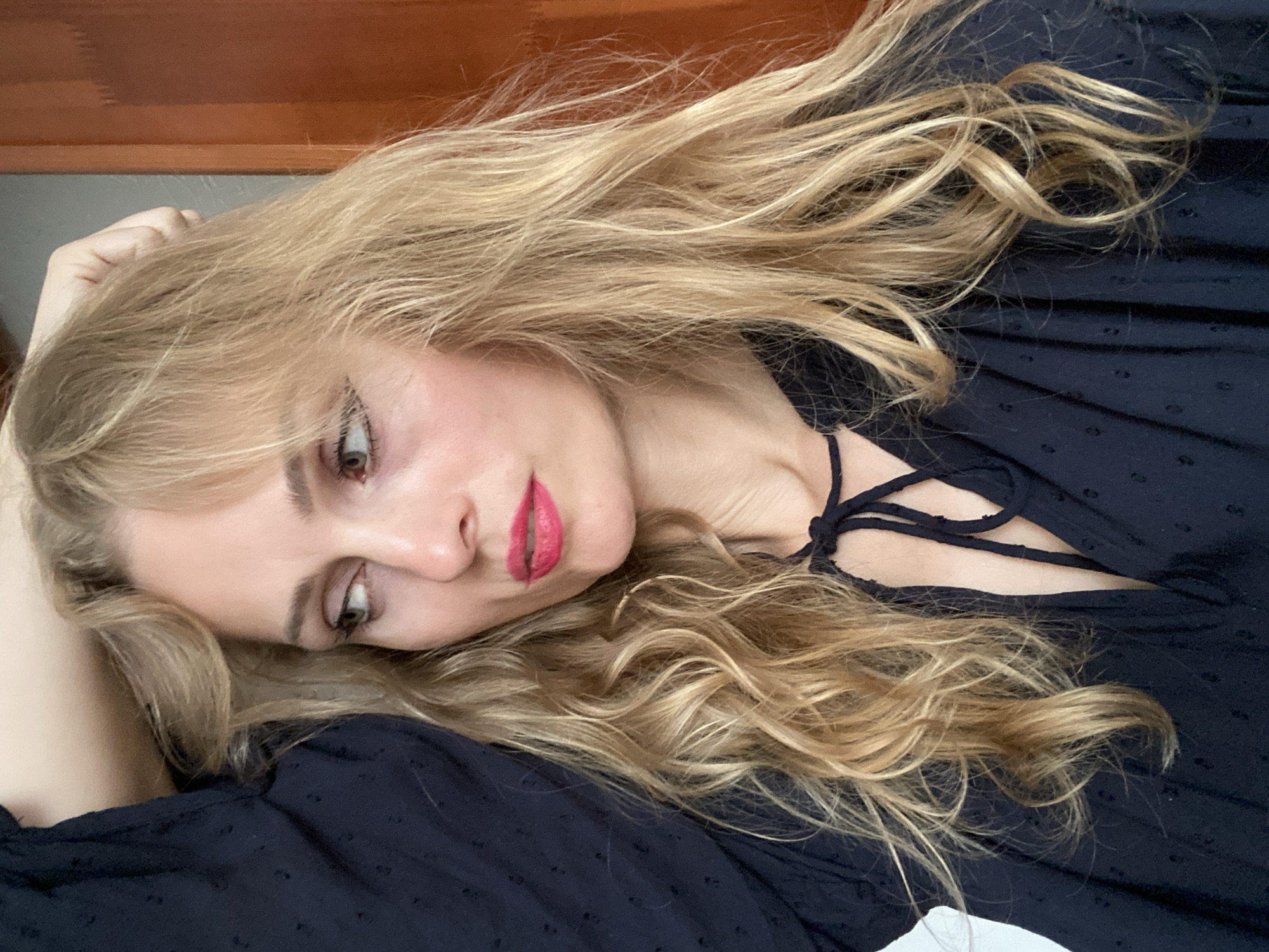 A woman with blonde, wavy hair and bright pink lipstick looks to her left, her left arm raised with her hand on her head. She is wearing a dark blue dress with a tie at the neckline. The background is a wooden door. The image was taken in Lida, Belarus.