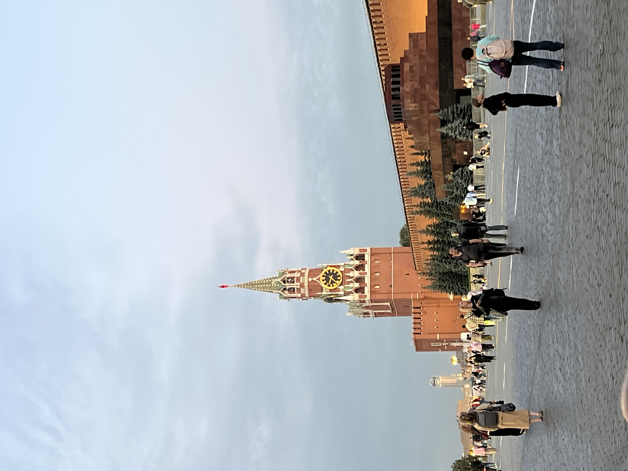 This image depicts a view of Red Square in Moscow, Russia, during a daytime with overcast skies. Dominating the frame is the Spasskaya Tower, a prominent landmark with a red facade, green tiered roof, and a large clock face with golden Roman numerals. To the right of the tower is a section of the red brick Kremlin wall, illuminated by warm light. In front of the wall, several dark green evergreen trees are visible.
The foreground of the image shows a cobblestone plaza, where numerous people are walking and standing. The individuals are varied in age and attire, and many are carrying bags or backpacks. Some are engaged in conversation, while others are looking around or at their phones. The scene suggests a public gathering or a tourist area. In the far distance, a tall modern building with a slender profile is visible against the lighter sky. The overall impression is that of a busy public space in a significant historical location.