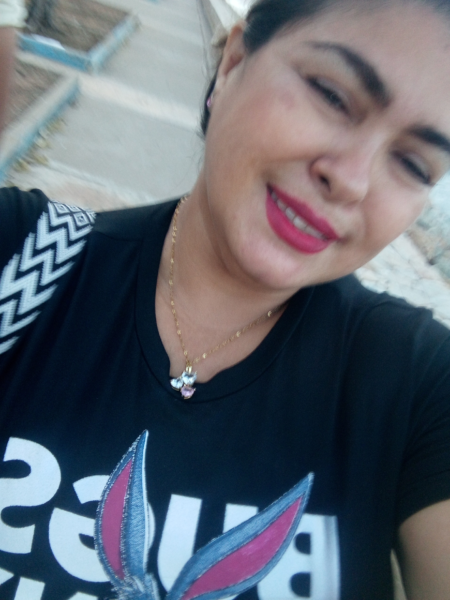 A woman is captured in a selfie, smiling and looking to the side with her eyes slightly closed. She is wearing a black t-shirt with a Bugs Bunny design on it. A gold chain with a gem pendant hangs around her neck. She appears to be outdoors in a city, the exact location of which is Unknown, Unknown.