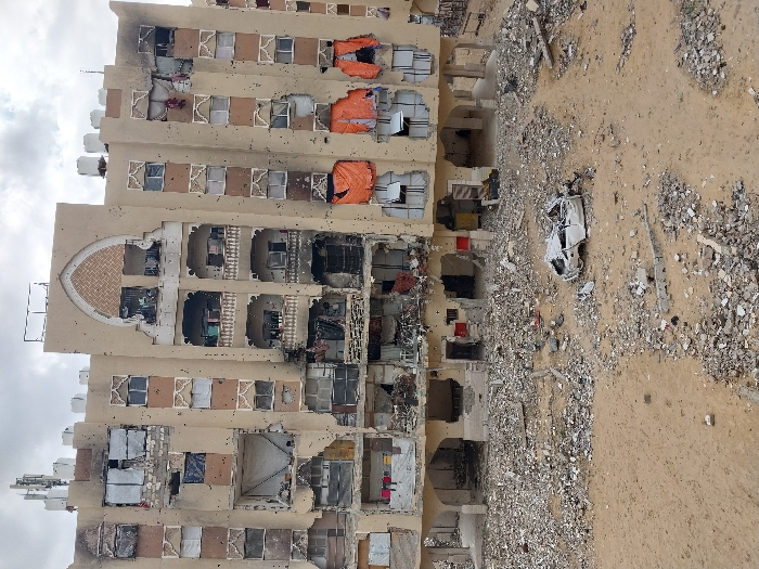 This image depicts a scene of widespread destruction in Khan Yunis, Palestinian Territory, under an overcast sky. The focal point is a multi-story residential building, light-tan in color, exhibiting extensive damage. The building's facade is riddled with numerous impact marks, and large sections of its outer walls are missing, exposing the derelict interiors of multiple apartments. Many windows are shattered or completely absent, with some openings covered by makeshift materials like orange and white tarpaulins or fabric. Signs of fire damage, including blackened walls and scorched remnants, are visible in various sections of the building. Decorative architectural elements, such as an arched motif near the top center and ornate balcony railings, are also present but show damage. Communication antennae and water tanks are visible on the rooftop. The ground in front of the building consists of sandy earth heavily covered with a dense layer of concrete rubble, fragmented building materials, and twisted metal debris. A severely damaged, white passenger vehicle, reduced to a crumpled shell, rests on this debris-strewn ground in the foreground. No individuals are visible in the scene.